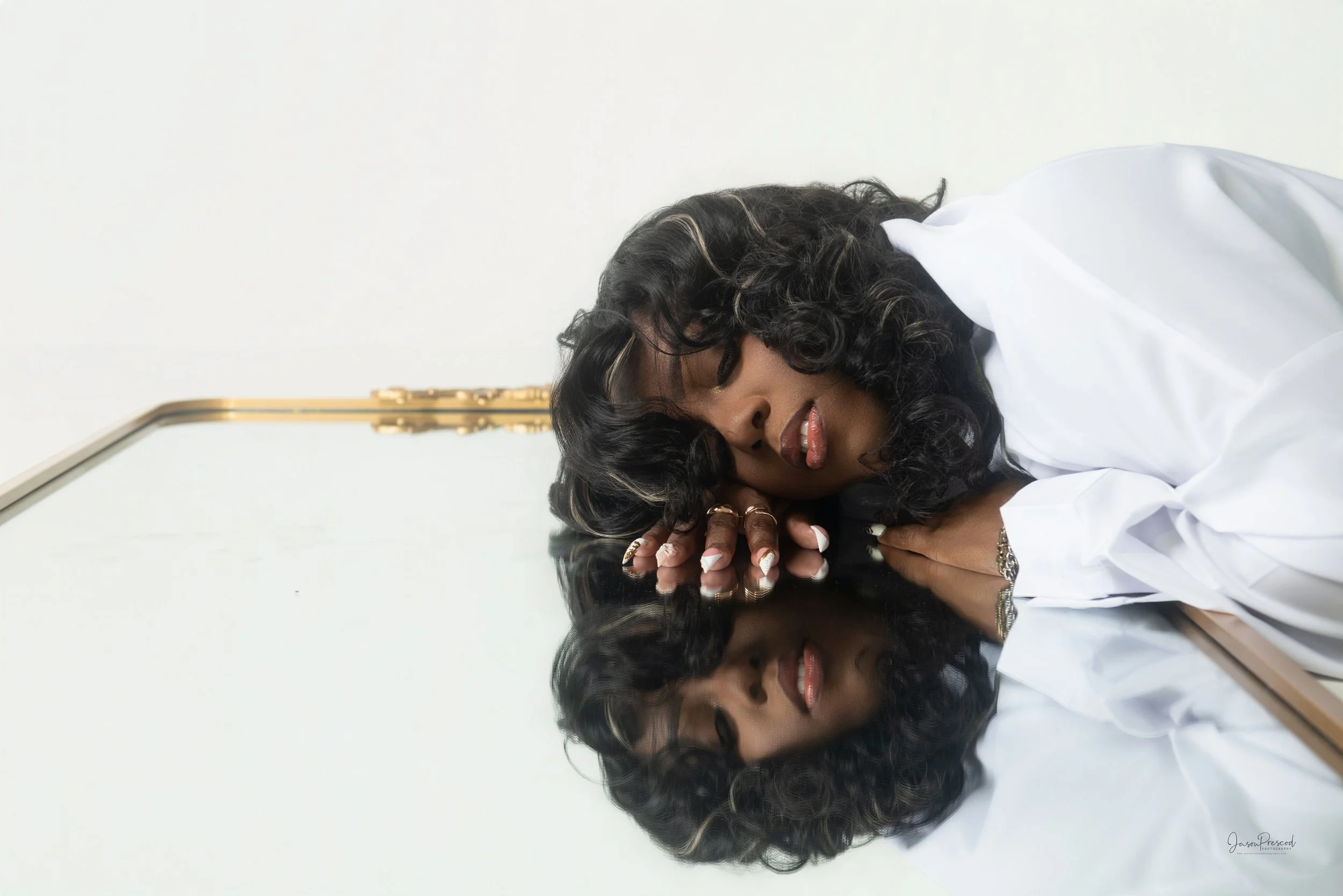 A woman with dark curly hair lying on a reflective black surface, resting her head on her folded arms, smiling with her eyes closed, wearing a white shirt against a plain white background.