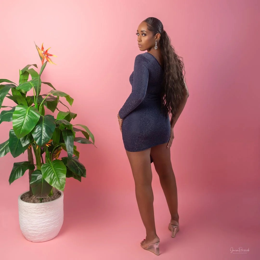 A woman with long, curly hair wearing a sparkly dark blue dress and clear high heels standing next to a large potted green plant against a pink background.