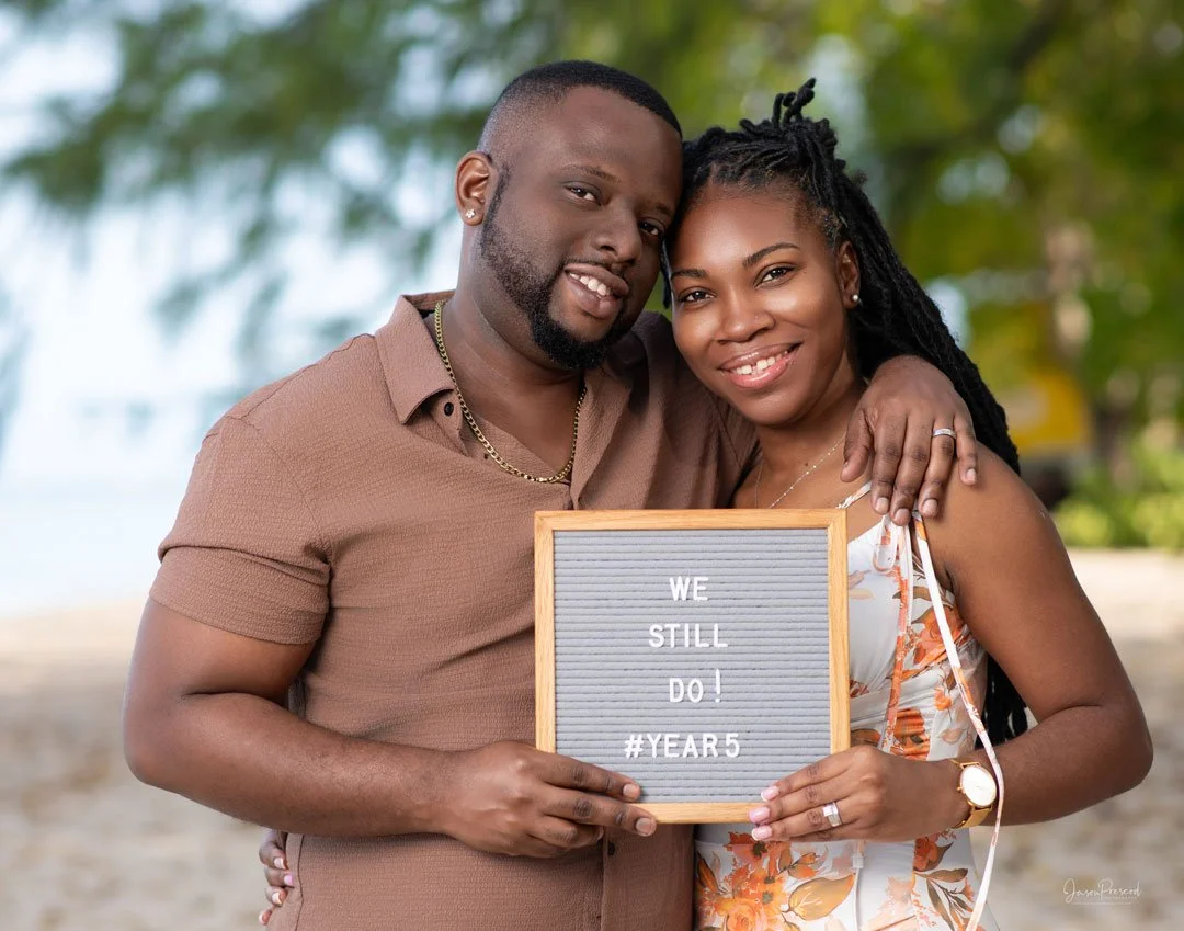 A smiling couple standing outdoors, holding a letter board that reads "WE STILL DO! #YEAR 5," celebrating their fifth wedding anniversary.