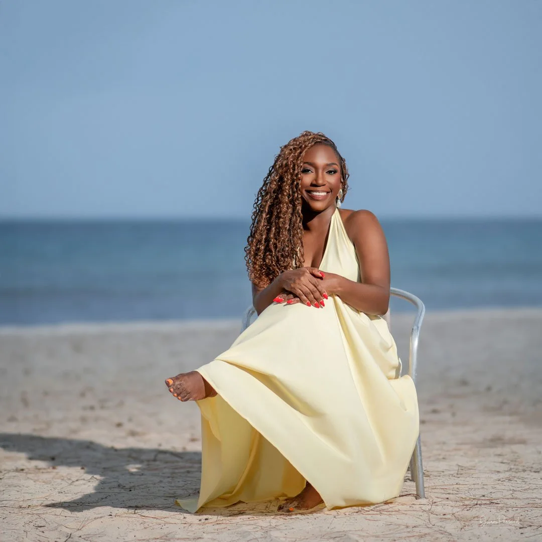 A woman with curly hair sitting on a chair at the beach, wearing a light yellow dress and smiling.