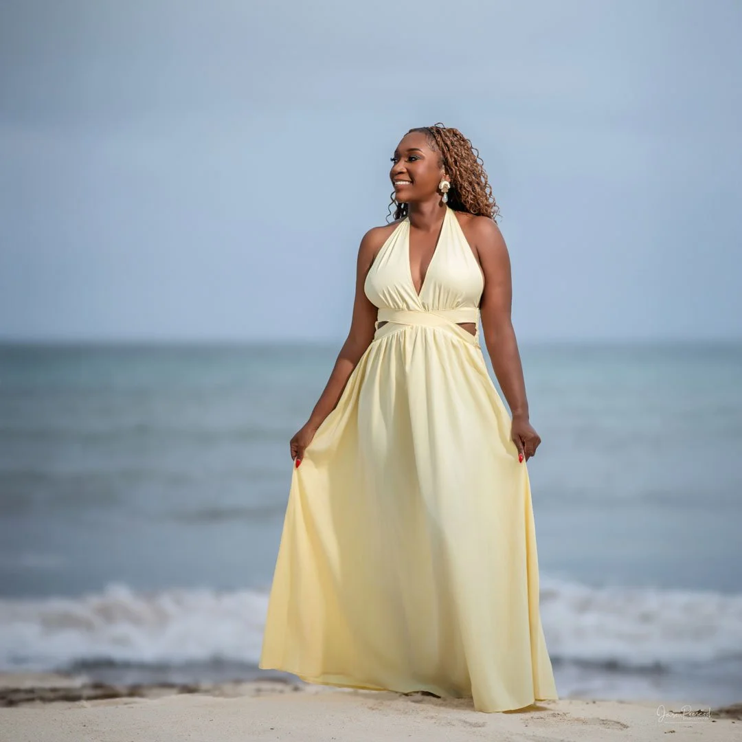 Woman in a yellow dress standing on the beach with the ocean in the background, smiling.