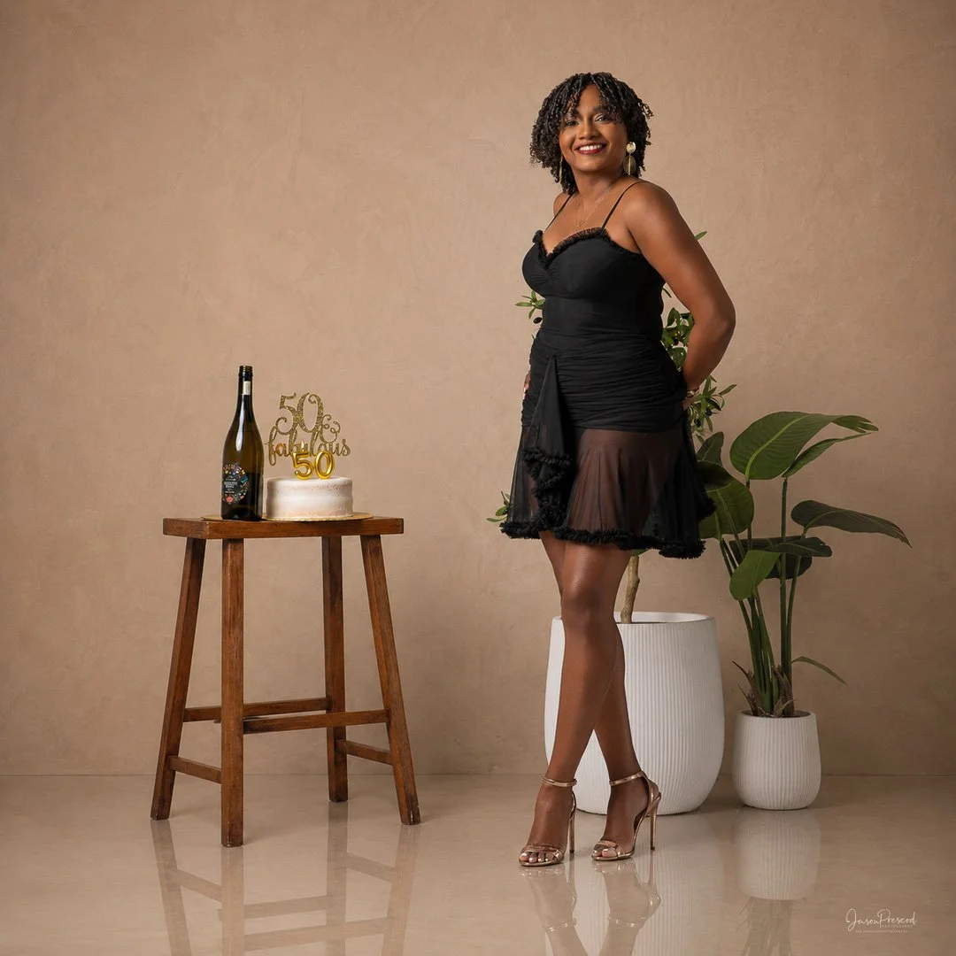 A woman in a black dress standing indoors with a celebration setup that includes a small cake, a bottle of champagne, and a golden '50 years' cake topper. There are two potted plants behind her.