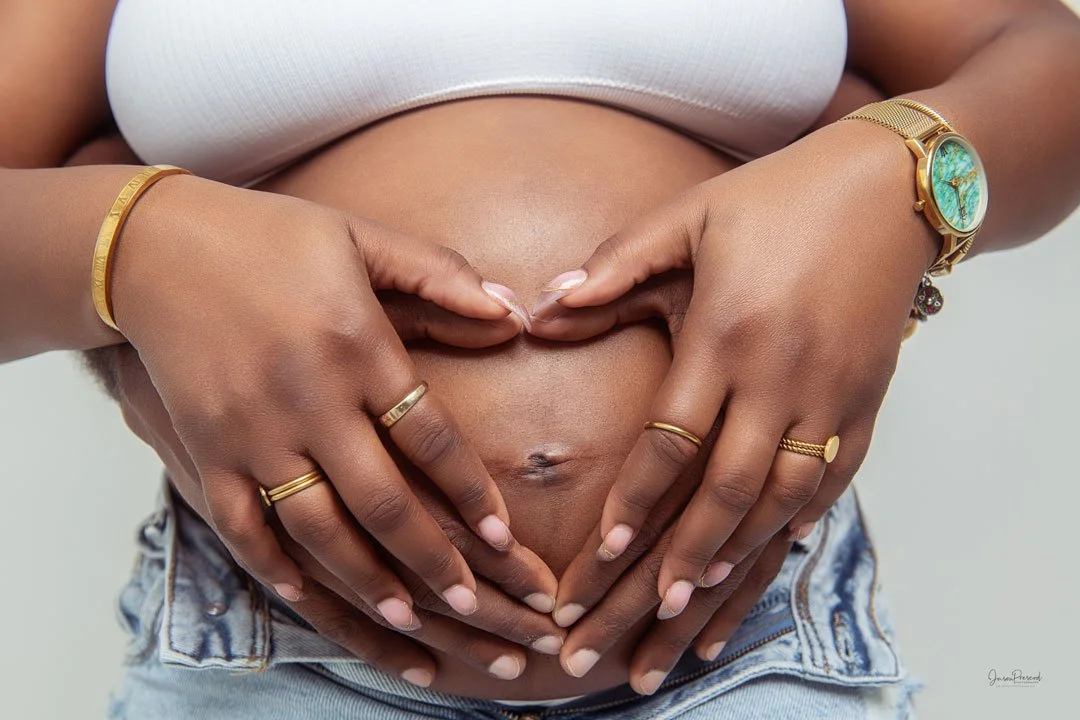 Close-up of a pregnant woman’s belly with several hands forming a heart shape around her stomach, wearing jewelry including rings, bracelets, and a watch.