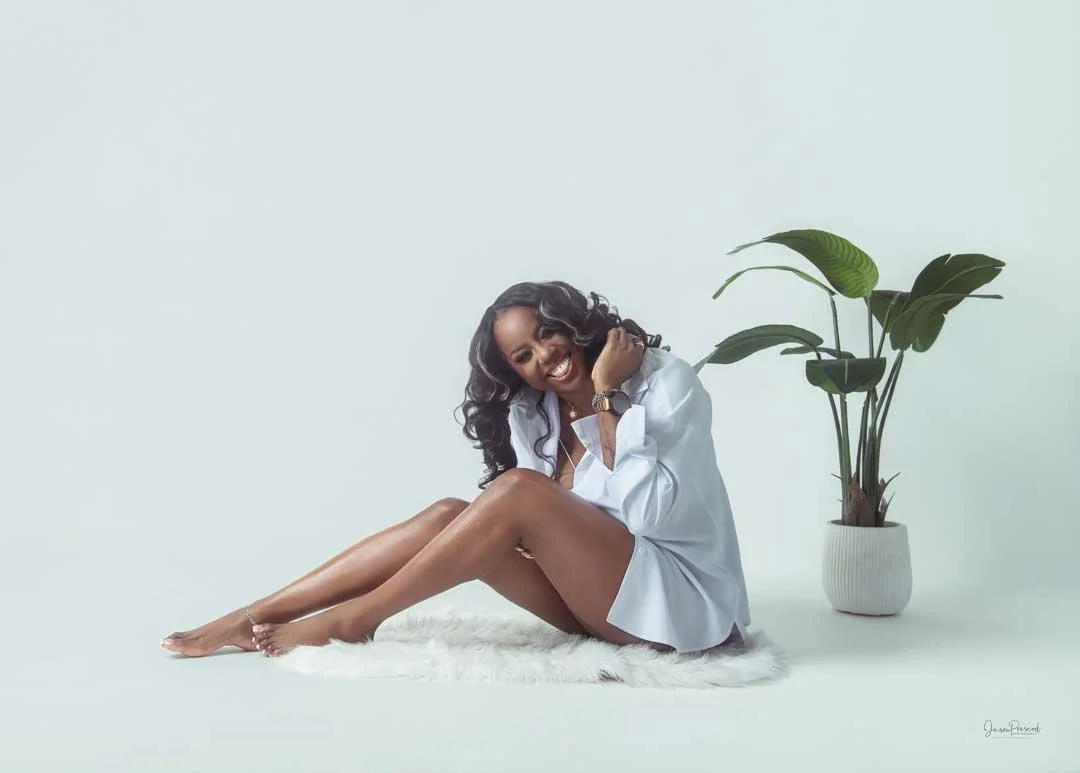 Smiling woman sitting on a white rug, wearing a white shirt, with a potted plant beside her, against a plain white background.