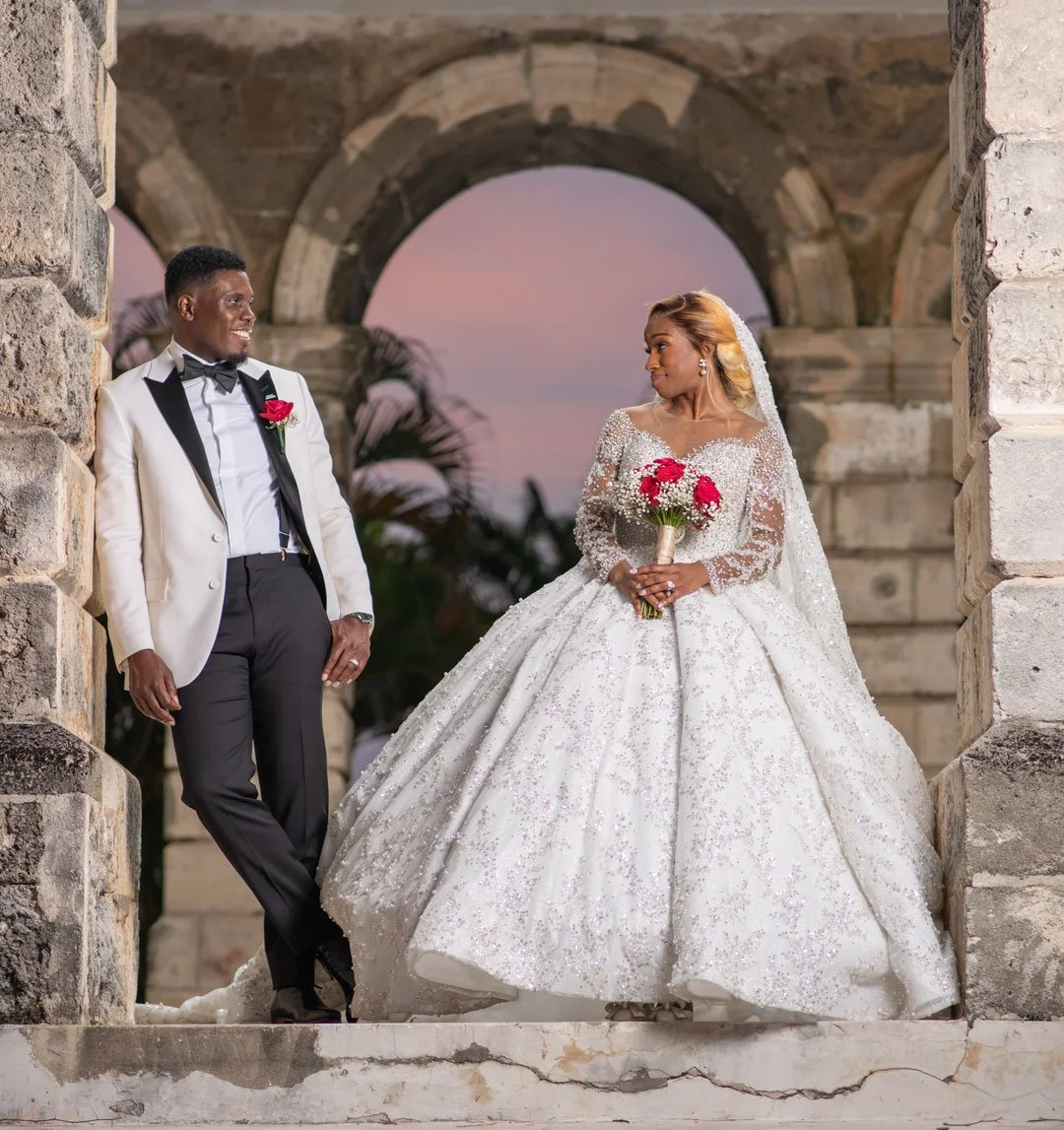 A newlywed couple stands under an ancient stone archway at sunset. The groom is in a white tuxedo with a black bow tie, and the bride is in a white, beaded wedding gown with a long veil, holding a bouquet of pink and red flowers. They are smiling at 