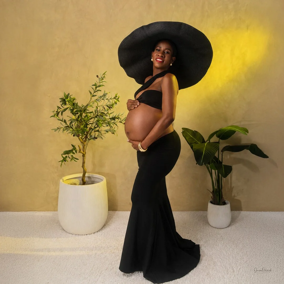 A pregnant woman in a black outfit and large black hat stands indoors, smiling while holding her belly, with two potted plants in the background and warm yellow lighting.