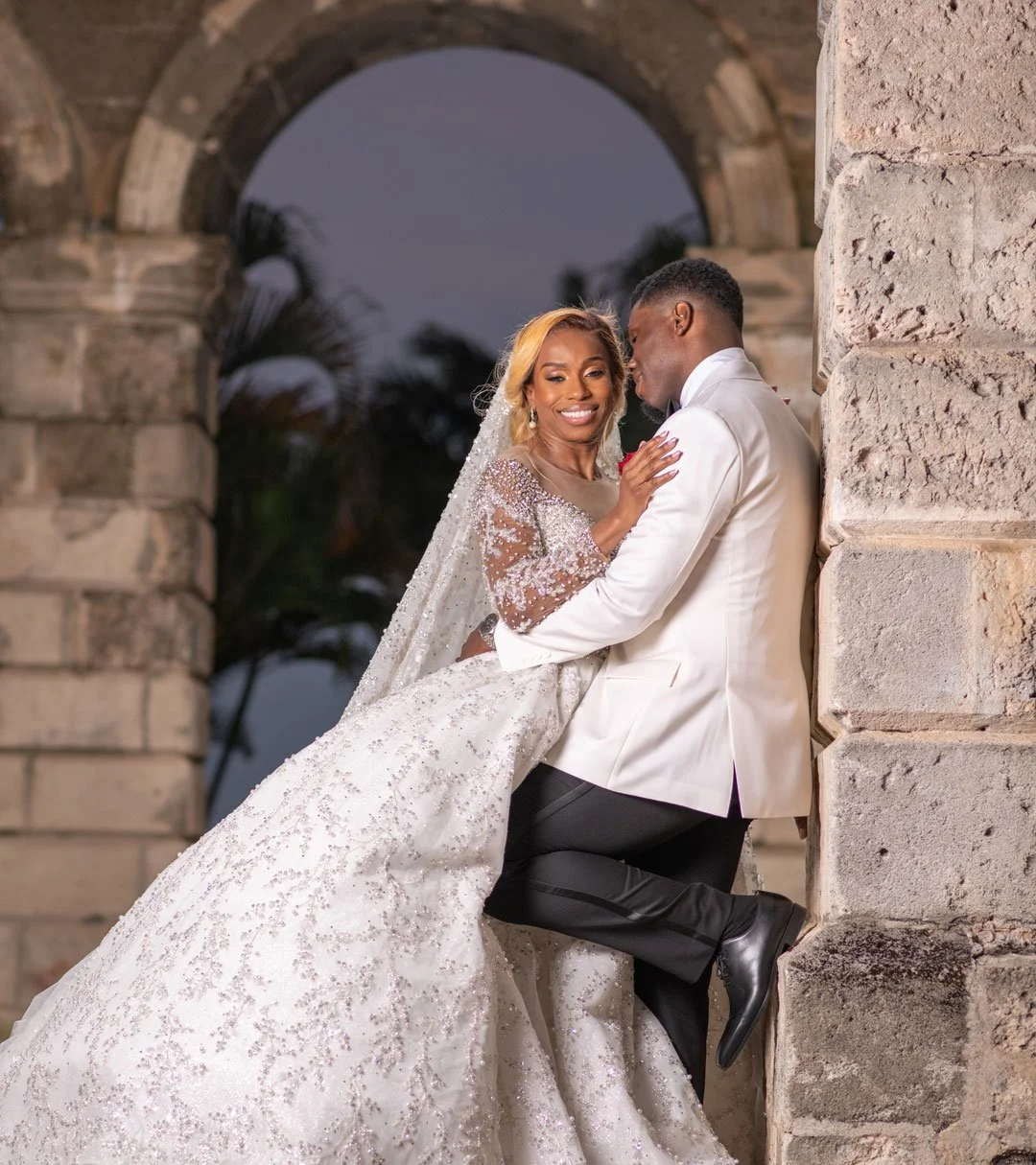 A bride and groom in wedding attire sharing a romantic moment outside, with the bride smiling and the groom leaning in for a kiss, framed by a stone archway at dusk.