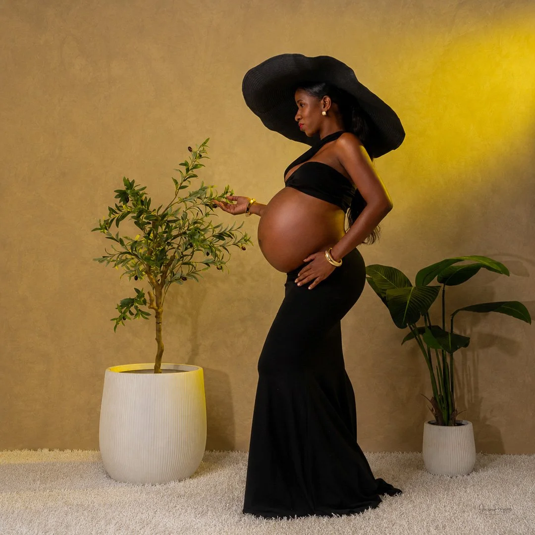 A pregnant woman wearing a black dress and wide-brimmed black hat standing indoors. She is holding a small branch with leaves, with potted plants nearby against a beige wall.