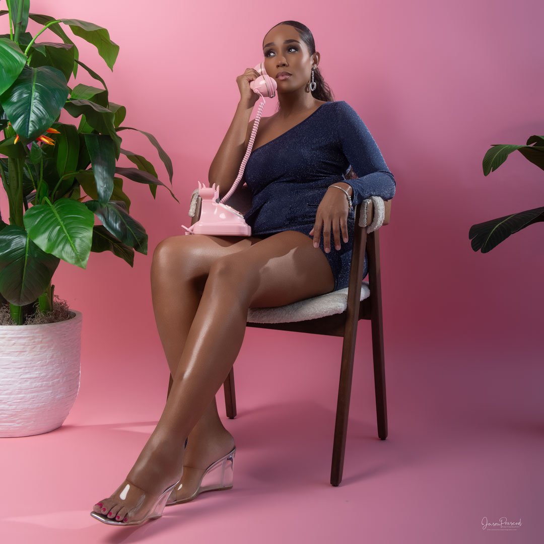 A woman in a sparkly navy dress with clear high-heeled shoes is sitting on a chair, talking on a pink vintage telephone, with a pink background and large green potted plants on either side.