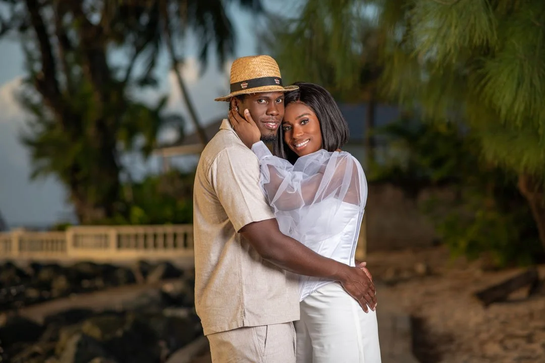 A couple embracing outdoors, with the man wearing a beige shirt and straw hat, and the woman in a white dress with sheer puffed sleeves, standing near greenery and a beach with rocks.