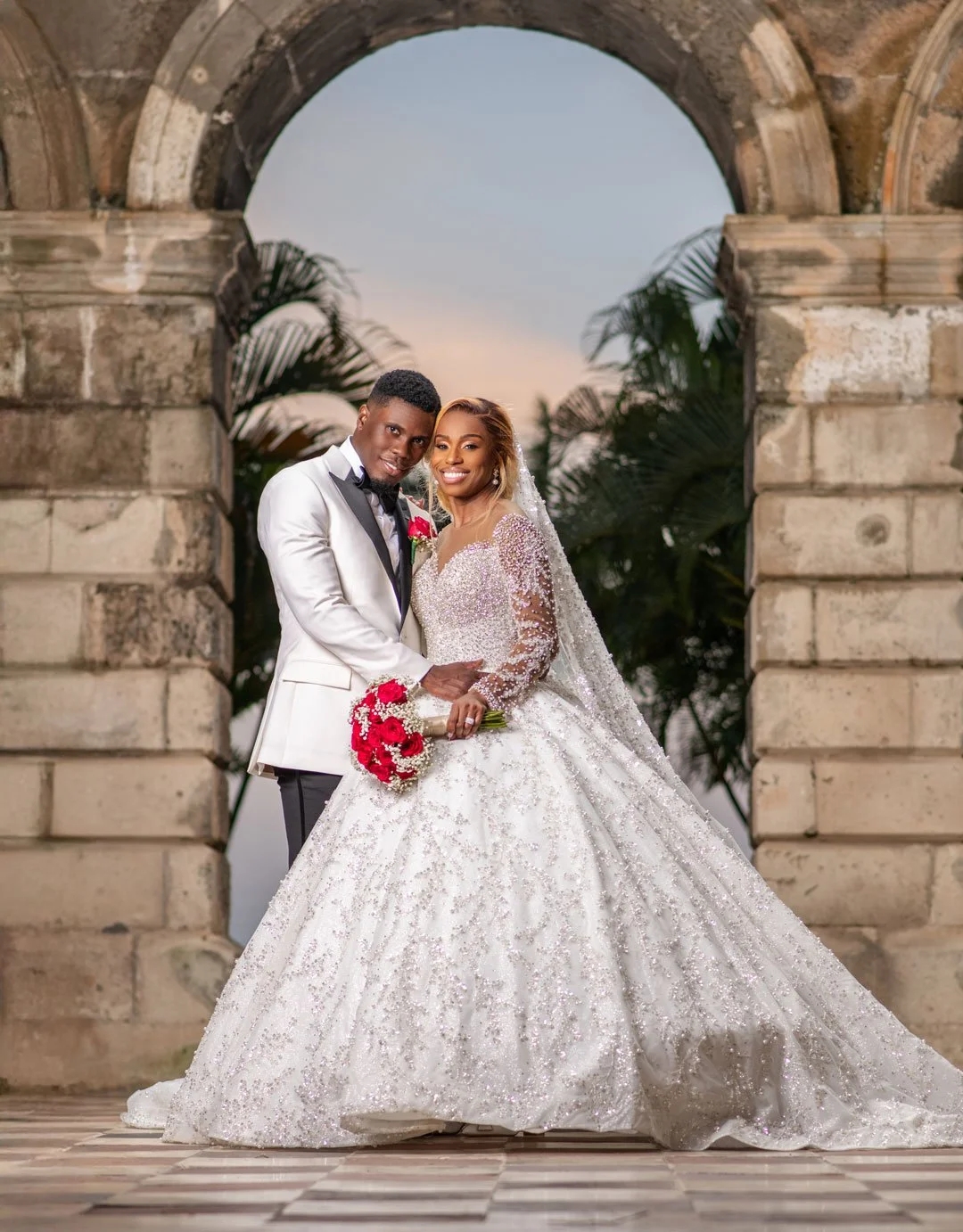 A bride and groom in wedding attire standing under a stone archway, with palm trees in the background, during sunset, celebrating their wedding.