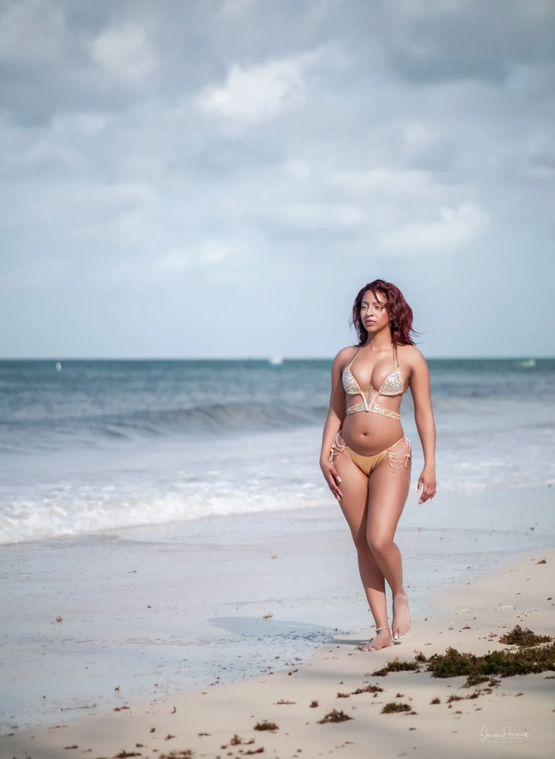 A woman with curly hair wearing a bikini walking along the sandy beach with the ocean and cloudy sky in the background.