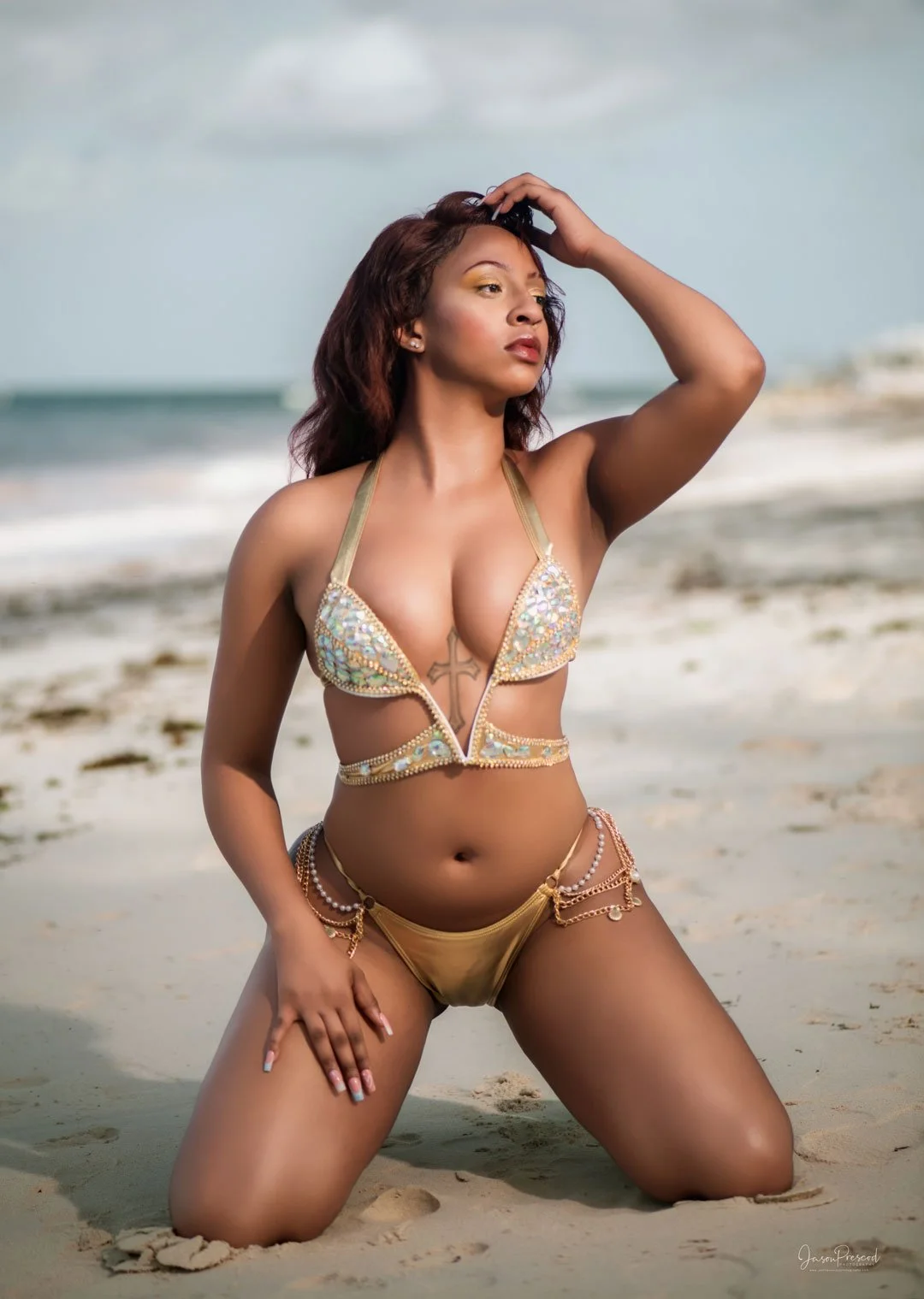 Woman in a embellished bikini posing on a sandy beach with ocean in background.