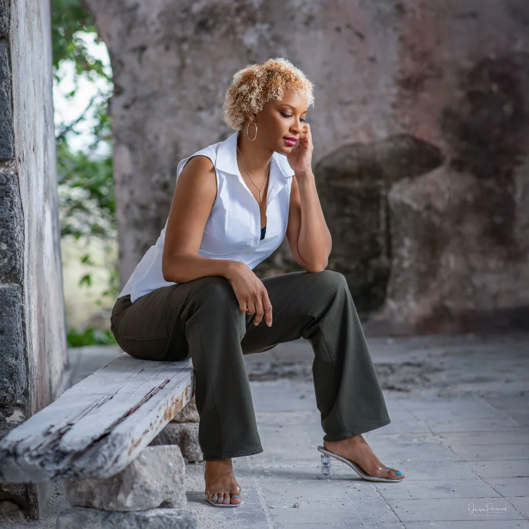A woman with curly blonde hair sitting on a weathered wooden bench against a stone wall, wearing a white sleeveless blouse, dark pants, and clear high-heeled shoes, with a contemplative expression.