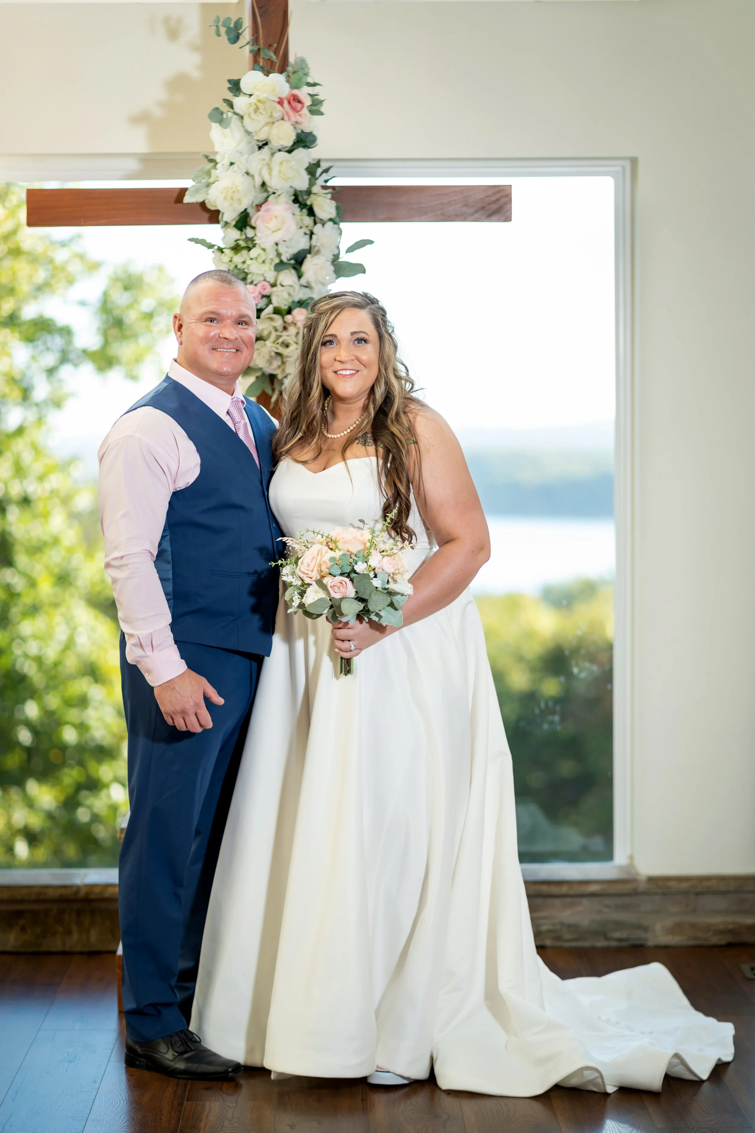 A newlywed couple standing inside near a window with a large wooden cross decorated with white and pink flowers behind them. The bride is holding a bouquet of flowers and wearing a white wedding dress, smiling. The groom is dressed in a blue vest and pants with a pink tie, also smiling.