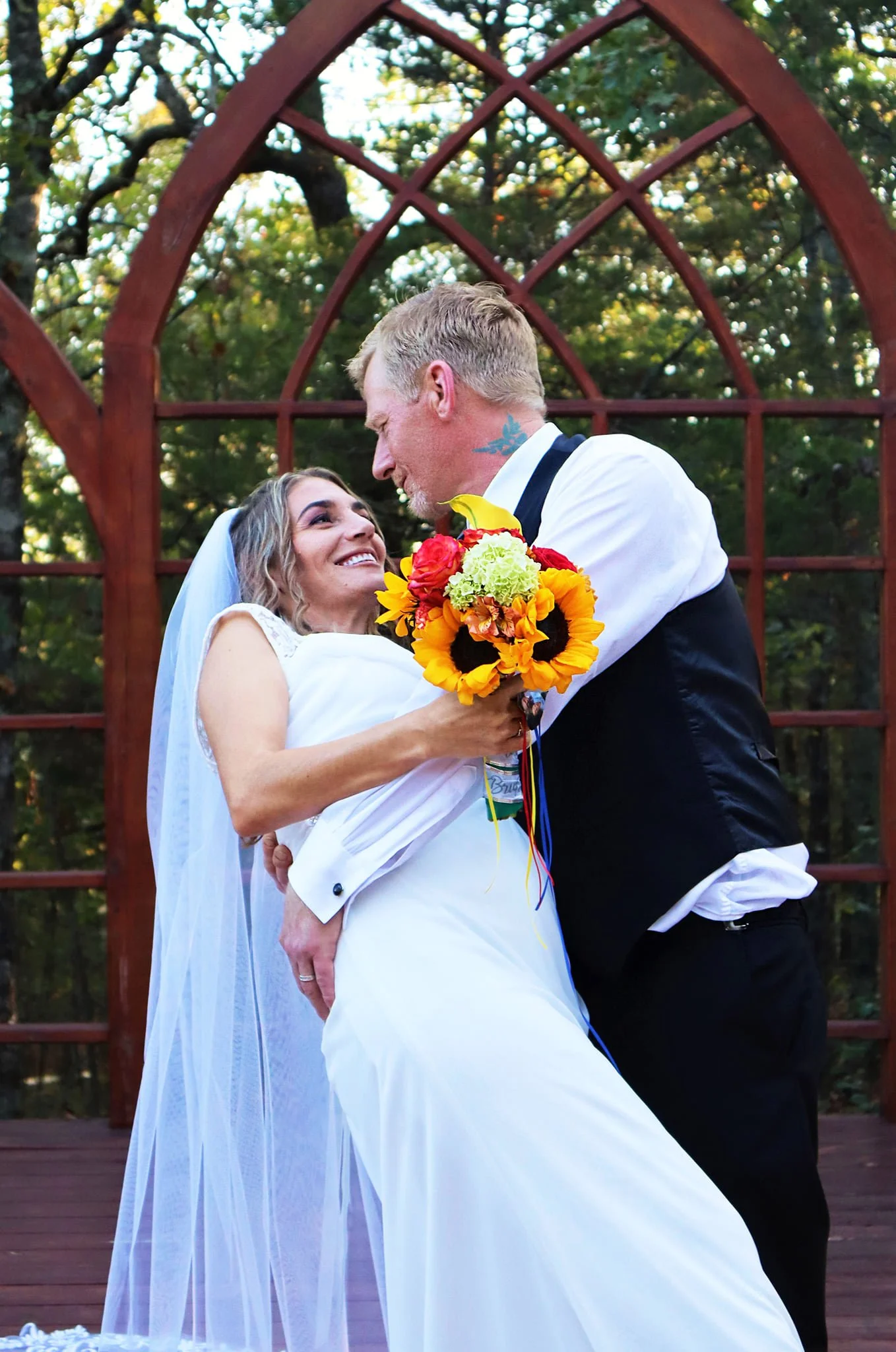 A bride and groom sharing a joyful moment during their wedding, with the bride holding a colorful bouquet of flowers and the groom leaning close to her, set against a backdrop of trees and a wooden structure.