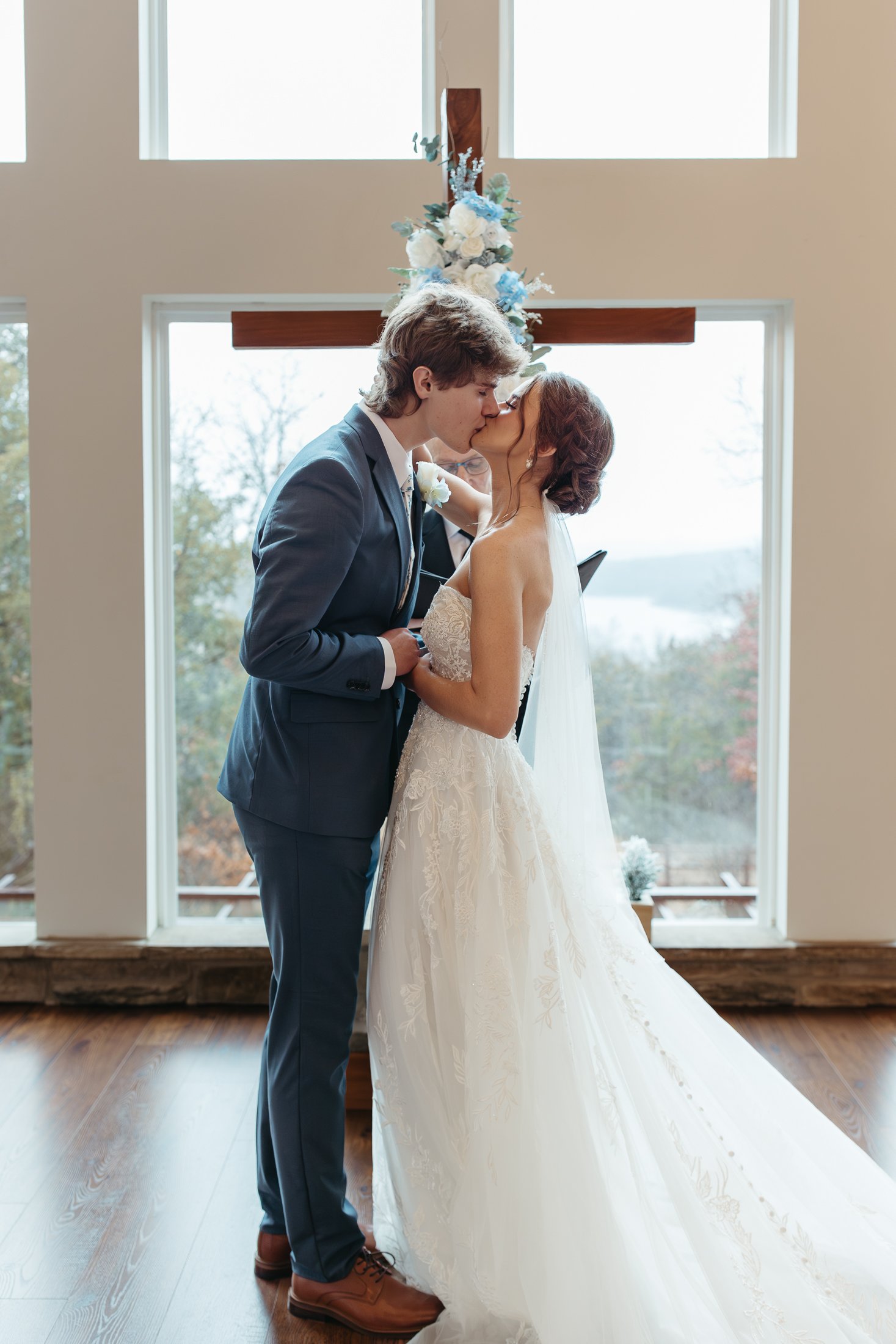 A couple in wedding attire sharing a kiss during their wedding ceremony inside a church with large windows and a cross with floral arrangements behind them.