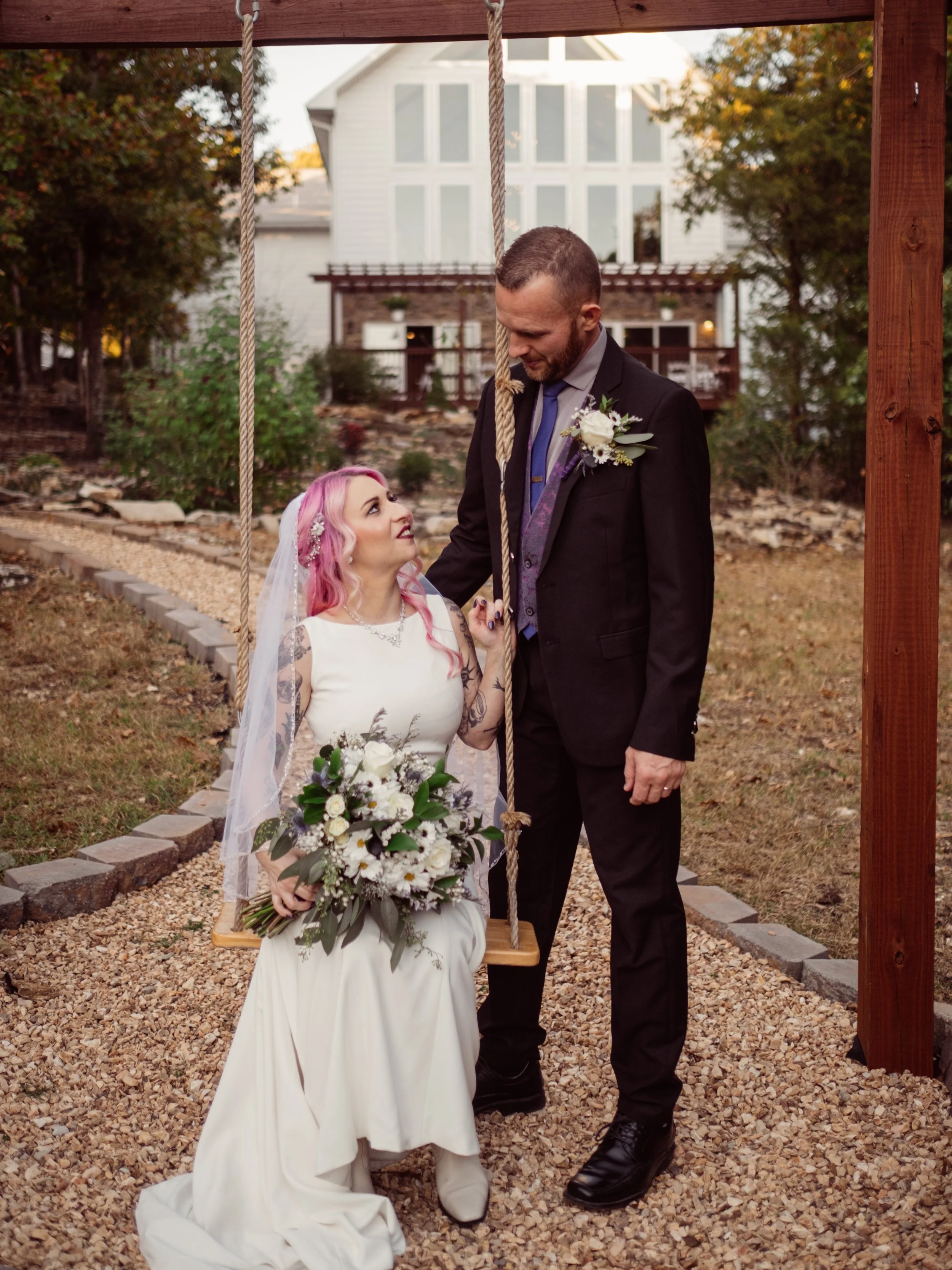 A bride with pink hair and tattoos, wearing a white dress and veil, holding a bouquet, sitting on a swing, and a groom in a dark suit with a boutonniere, standing close to her, holding her hand, outside in a garden area.