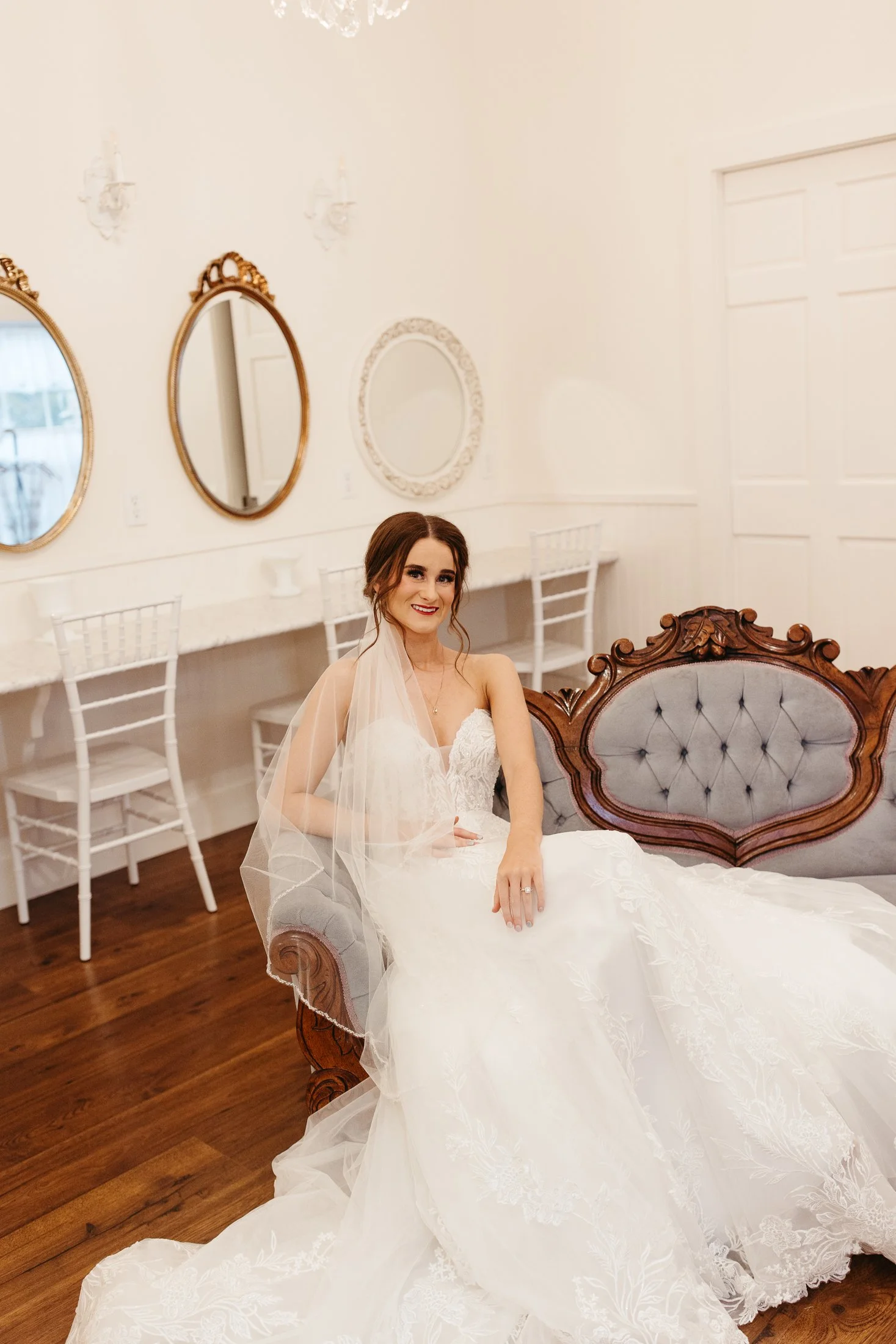 A bride wearing a white wedding dress with lace details, sitting on a vintage gray and wood sofa in a decorated room with white walls and chandeliers.