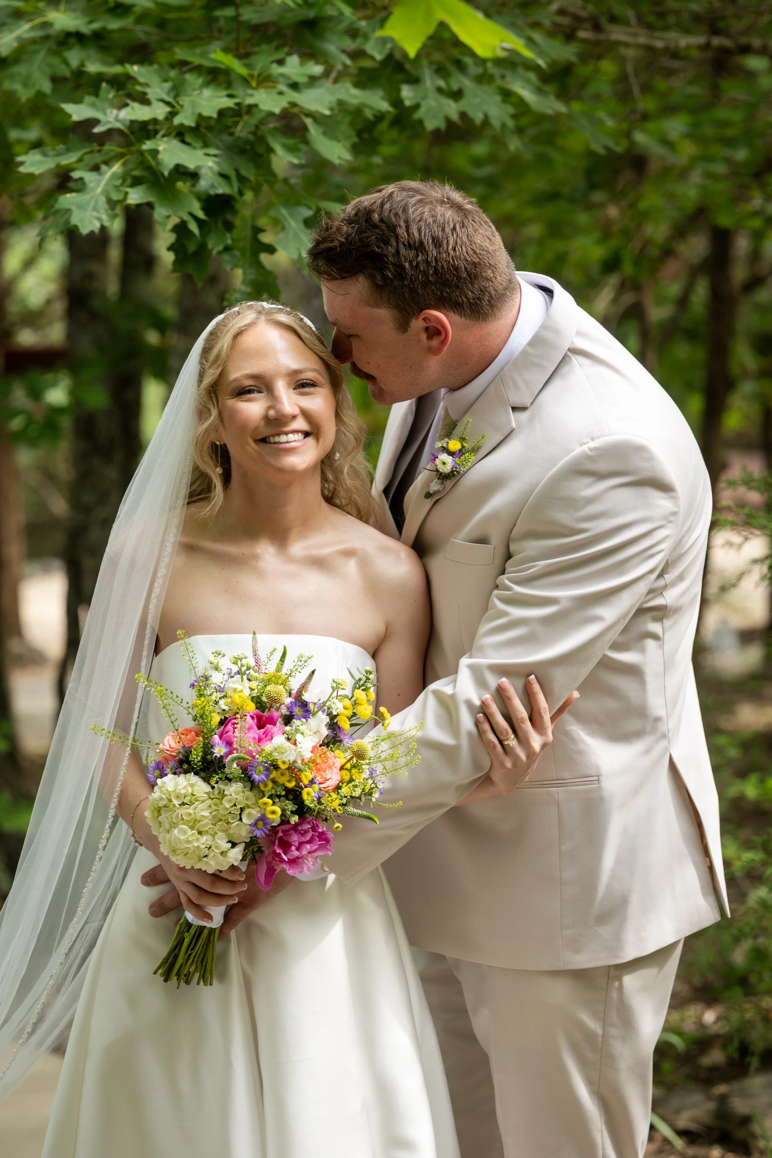 A bride and groom at their outdoor wedding, with the groom kissing the bride's forehead. The bride is smiling and holding a colorful bouquet of flowers. The groom is dressed in a beige suit with a boutonniere, and the bride is wearing a strapless white wedding dress with a veil.