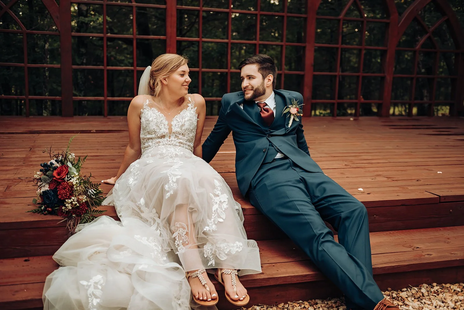 A bride and groom sitting on a wooden stage outdoors. The bride is in a white lace wedding dress with a veil, holding a bouquet of flowers. The groom is in a dark blue suit with a red tie and boutonniere. They are smiling at each other in a candid mo