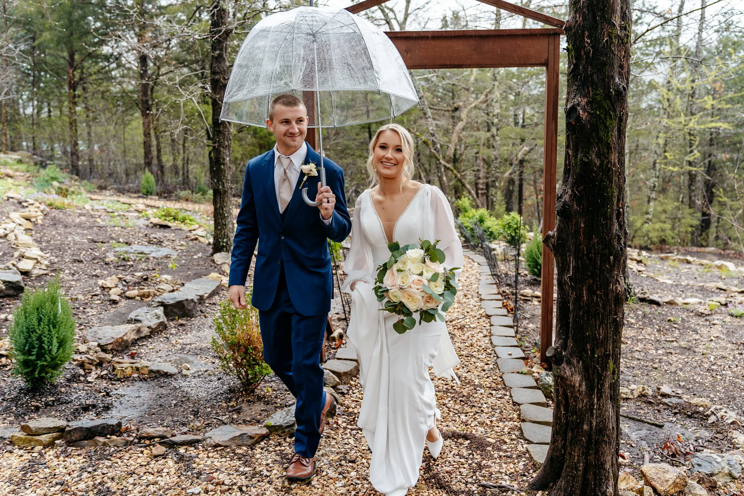 A newlywed couple walking outdoors in a wooded area, holding a transparent umbrella. The groom wears a blue suit and the bride wears a white wedding dress and holds a bouquet of white roses.