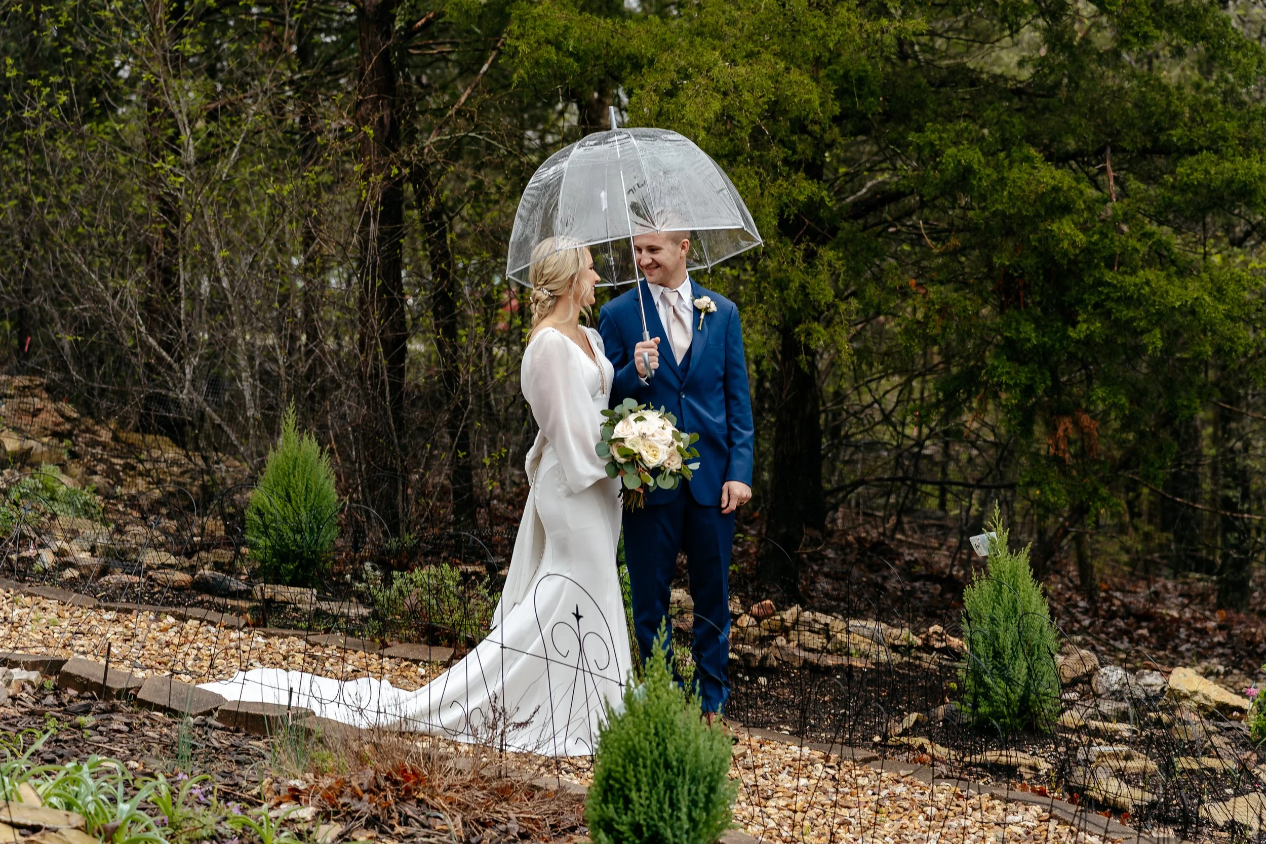 A bride and groom standing outdoors under a clear umbrella during a rainy wedding ceremony. The bride is holding a bouquet and wearing a white wedding gown, and the groom is wearing a blue suit with a boutonniere.