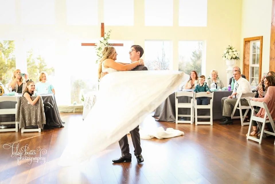 A bride and groom dancing during their wedding reception with guests seated at tables around them in a well-lit hall.