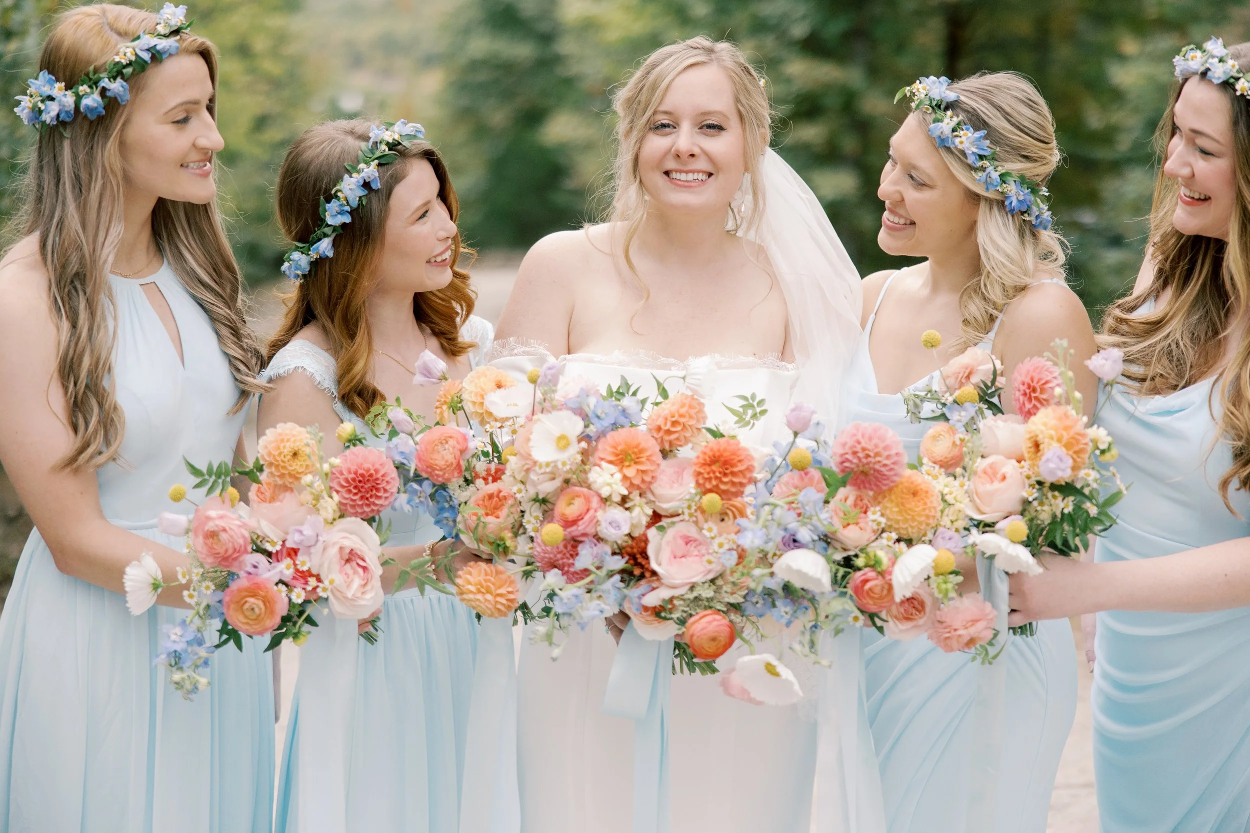 A bride in a white wedding dress smiling, surrounded by five bridesmaids in sky blue dresses wearing floral crowns made of blue and white flowers, and holding a large bouquet of multicolored flowers, outdoors with green trees in the background.