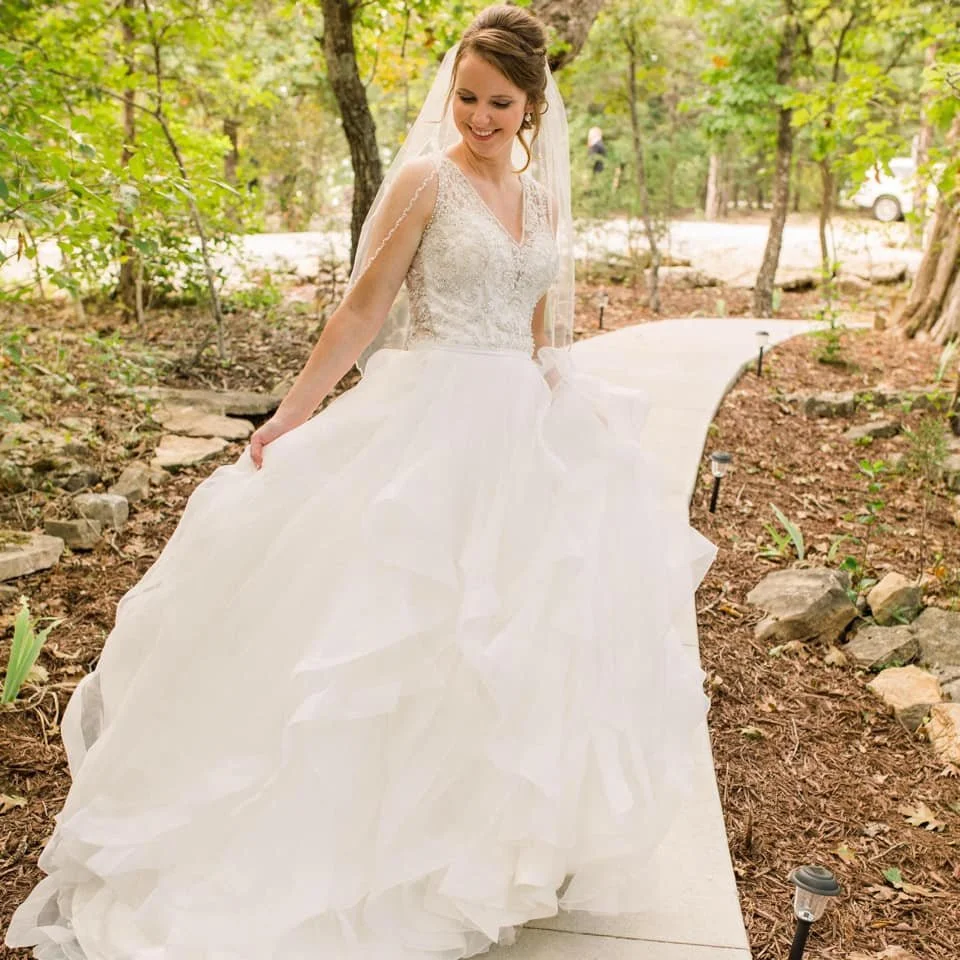 A bride in a white wedding dress walking on a forest path, holding the skirt of her dress and smiling.