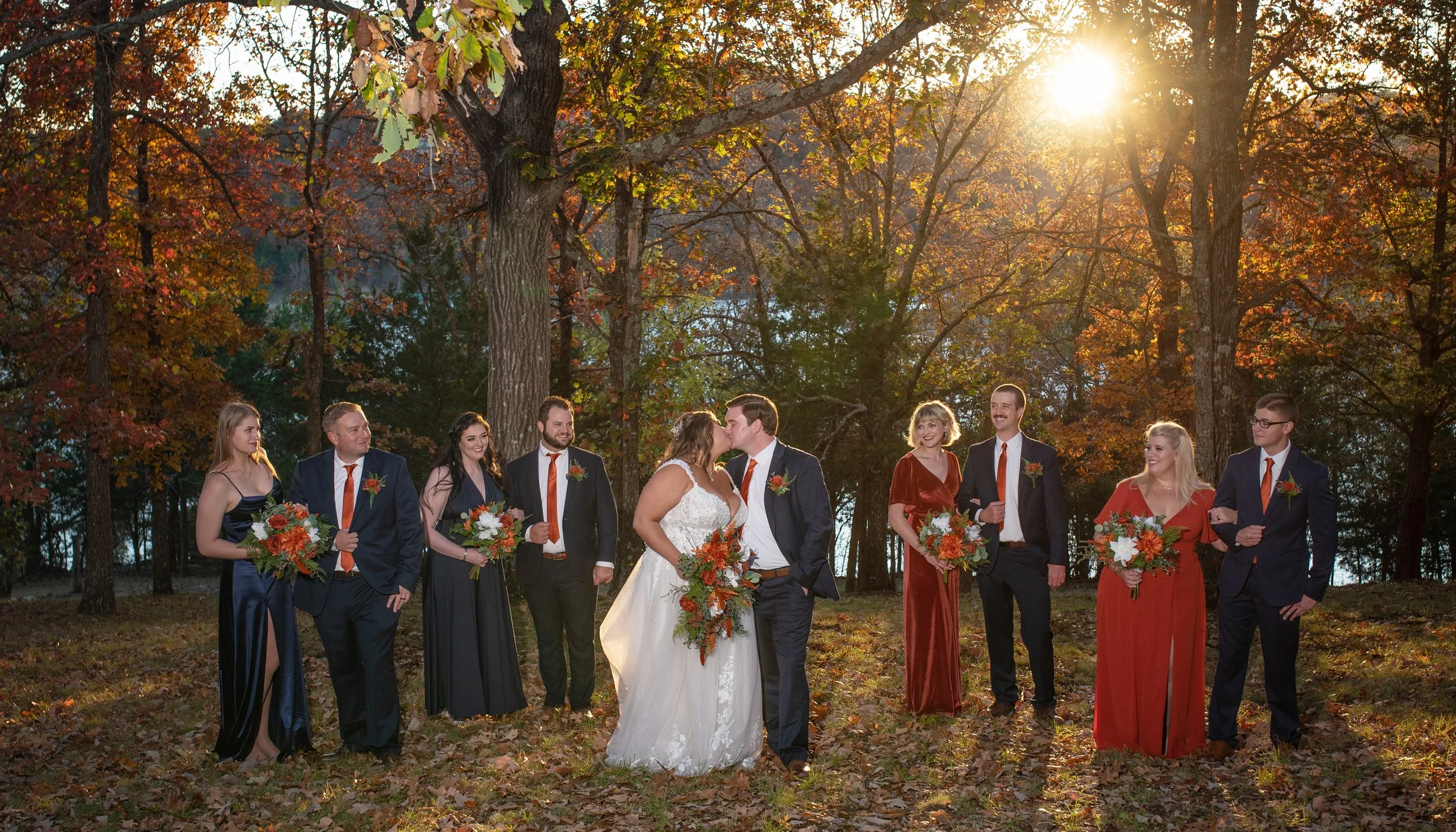 A group of people, including a bride and groom, standing outdoors in front of autumn trees, with the sun shining through the trees, during a wedding celebration.