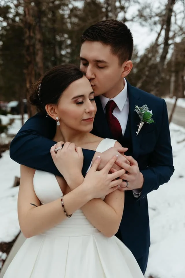 A bride and groom in wedding attire embrace outdoors in a snowy forested area, with the groom kissing the bride's forehead.