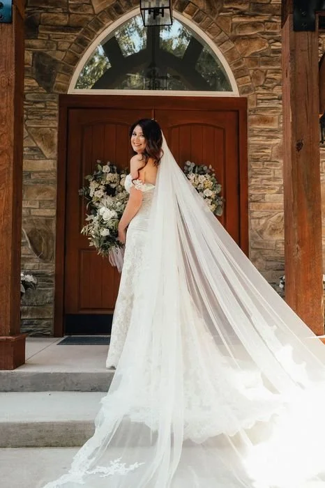 Bridal portrait of a bride in a white wedding dress with a long veil standing in front of a wooden door with floral arrangements on either side.