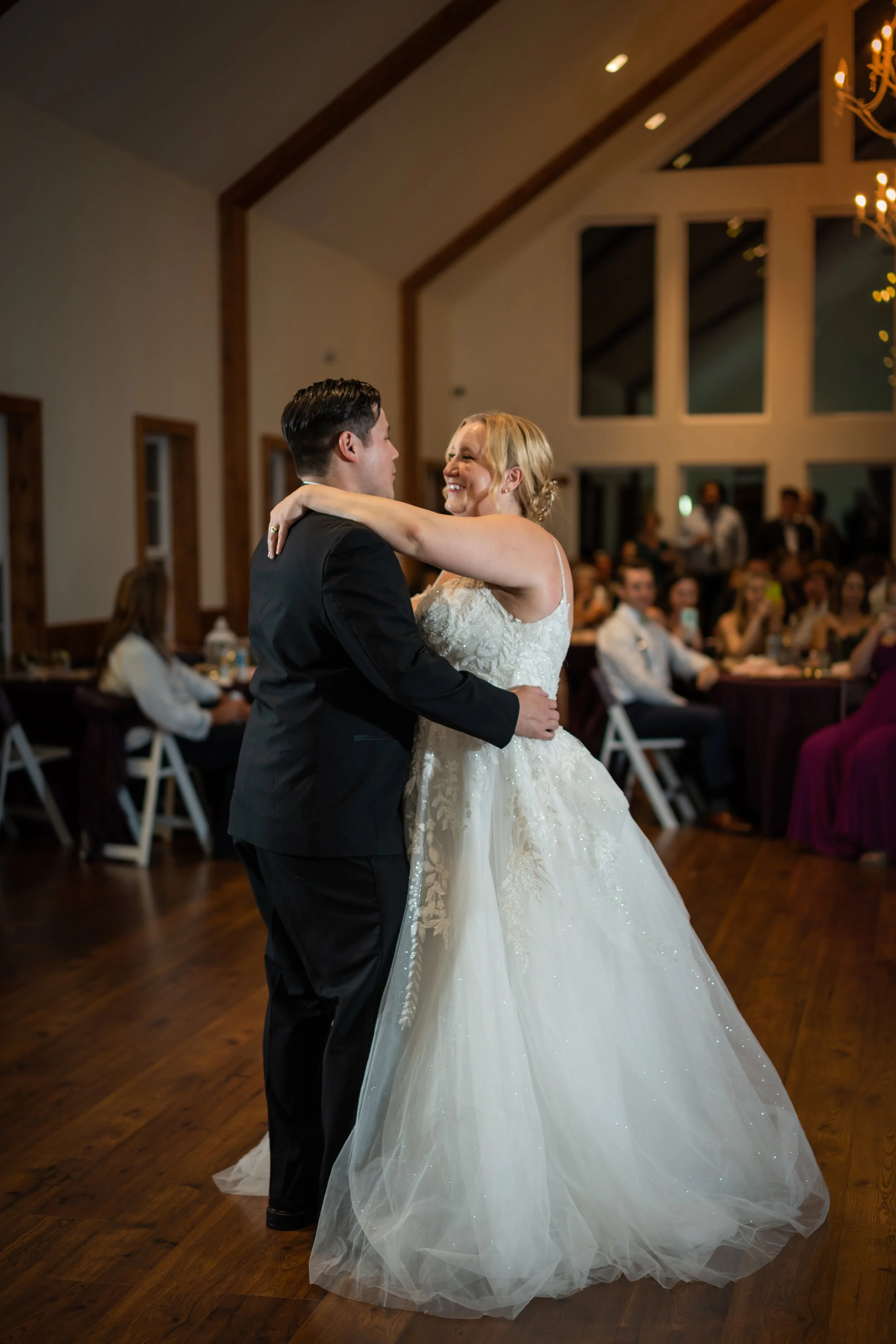 A bride and groom sharing their first dance at a wedding reception in a decorated hall with wooden beams and large windows.