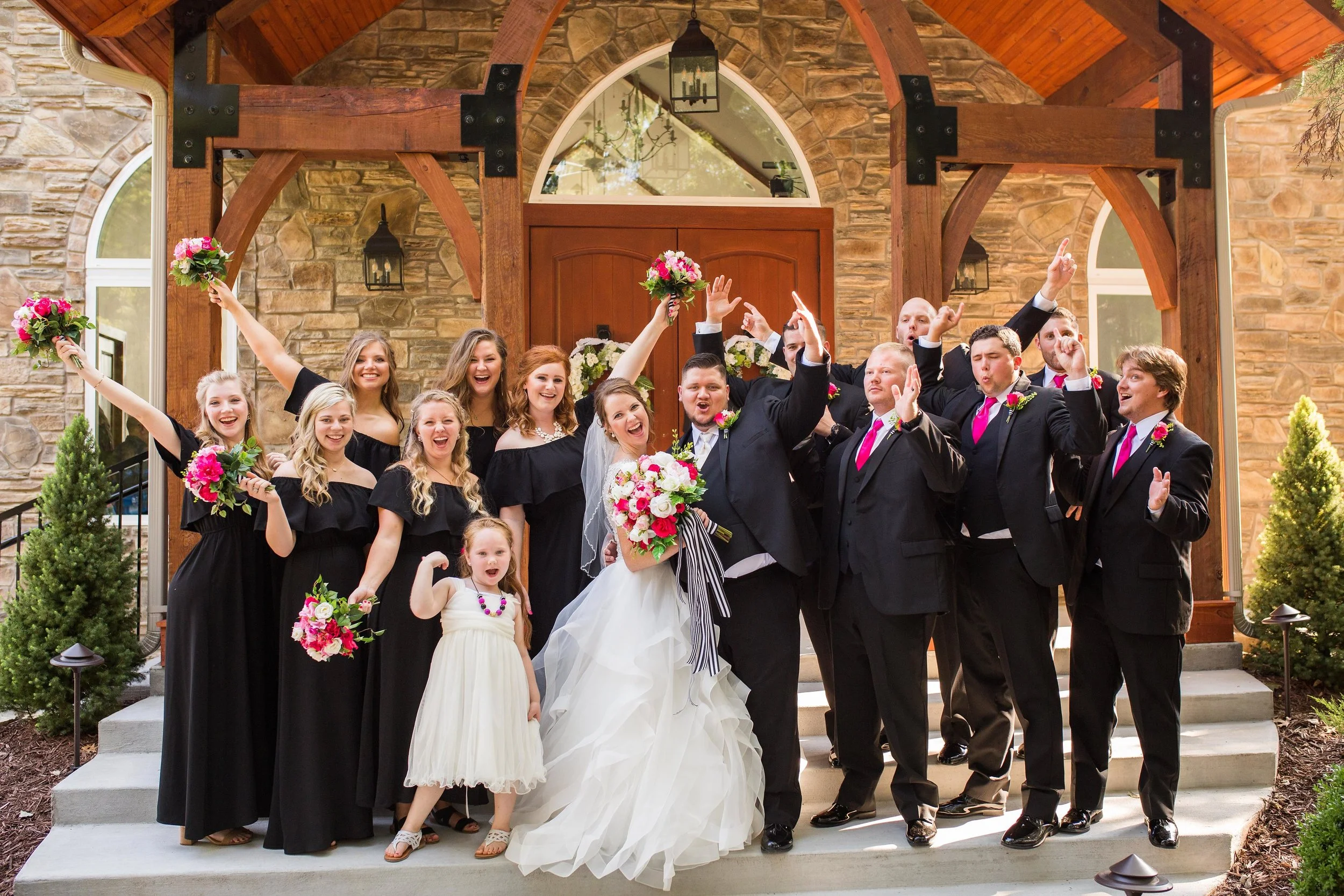 Group of people celebrating outside a house, with a bride and groom at the center. The bride is holding a bouquet, and everyone is smiling and making cheerful gestures. The house features stone walls, arched windows, and wooden accents.