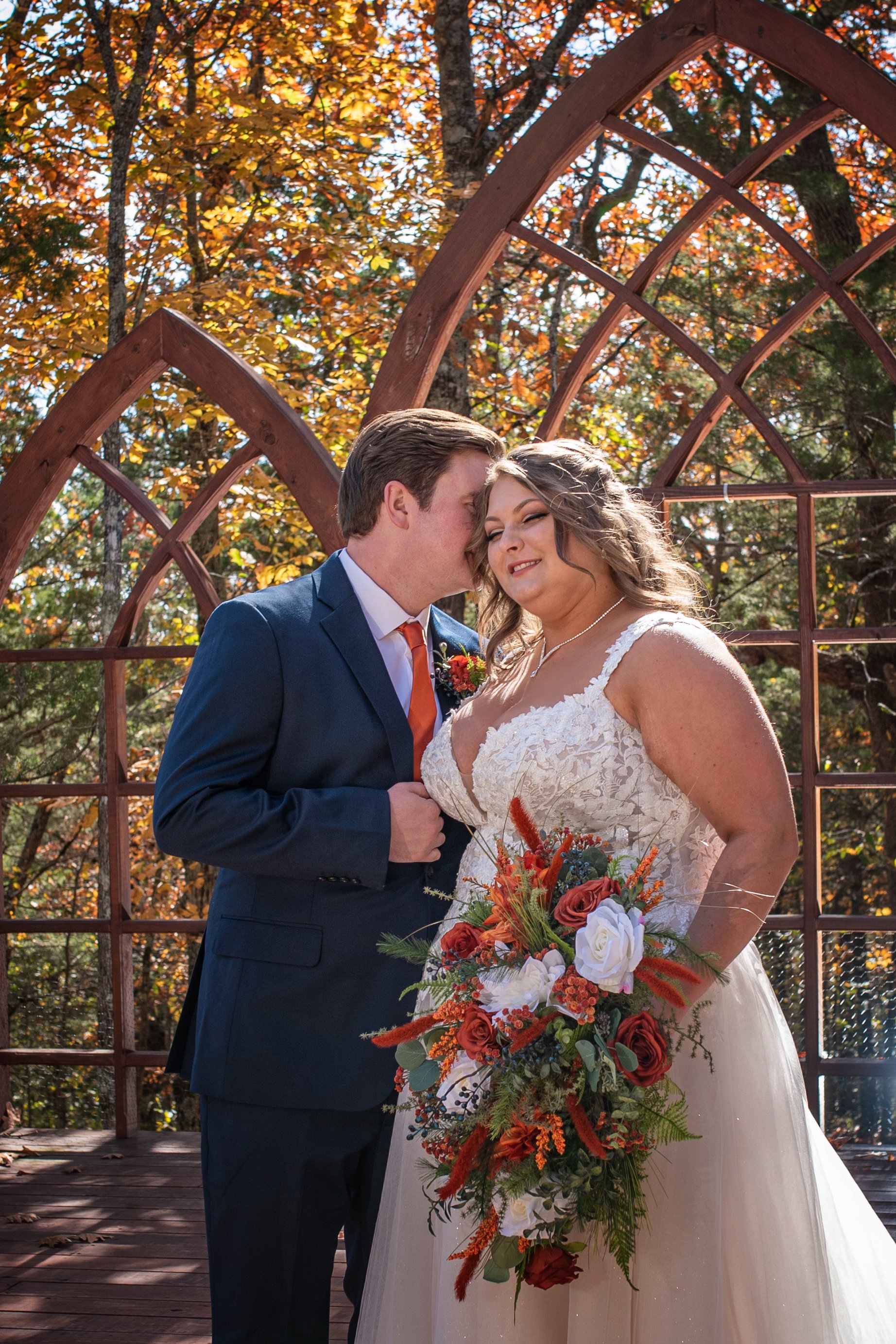 A bride and groom sharing a moment on their wedding day outdoors, with autumn trees in the background and a decorative wooden arch.
