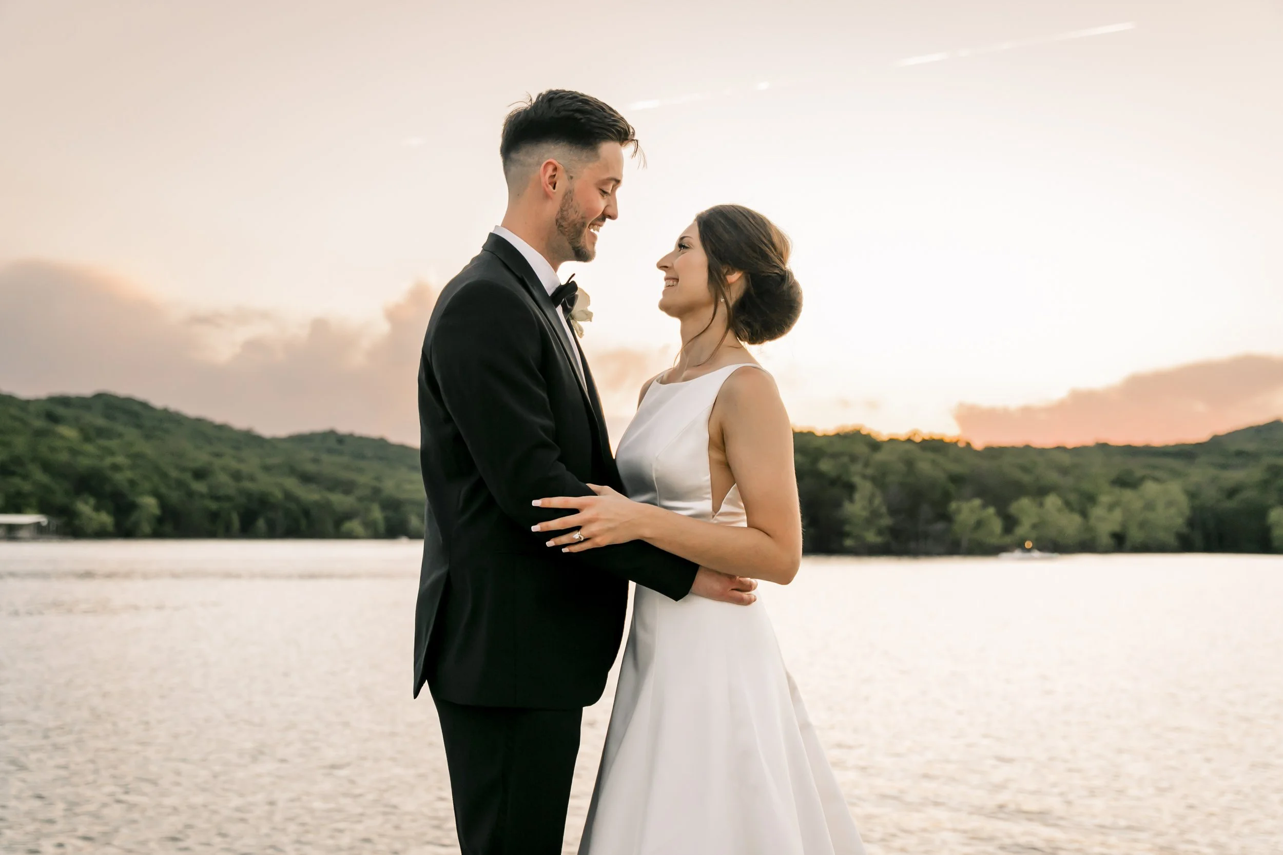 A newlywed couple in wedding attire holding each other by a body of water during sunset, with green hills in the background.