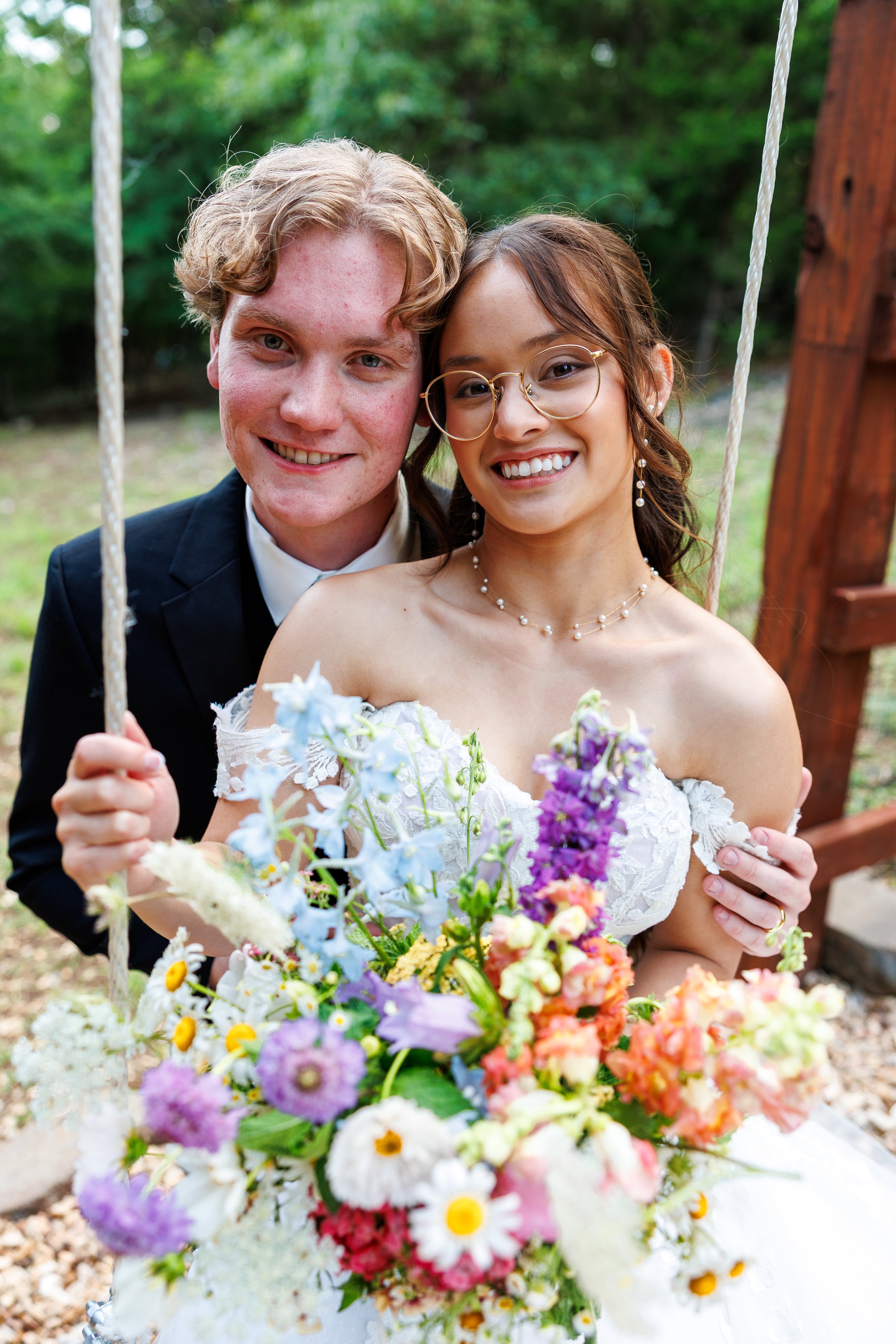 A smiling couple at a wedding outdoors, holding a swing, with the woman in a white off-shoulder wedding dress and the man in a dark suit, surrounded by colorful flowers.