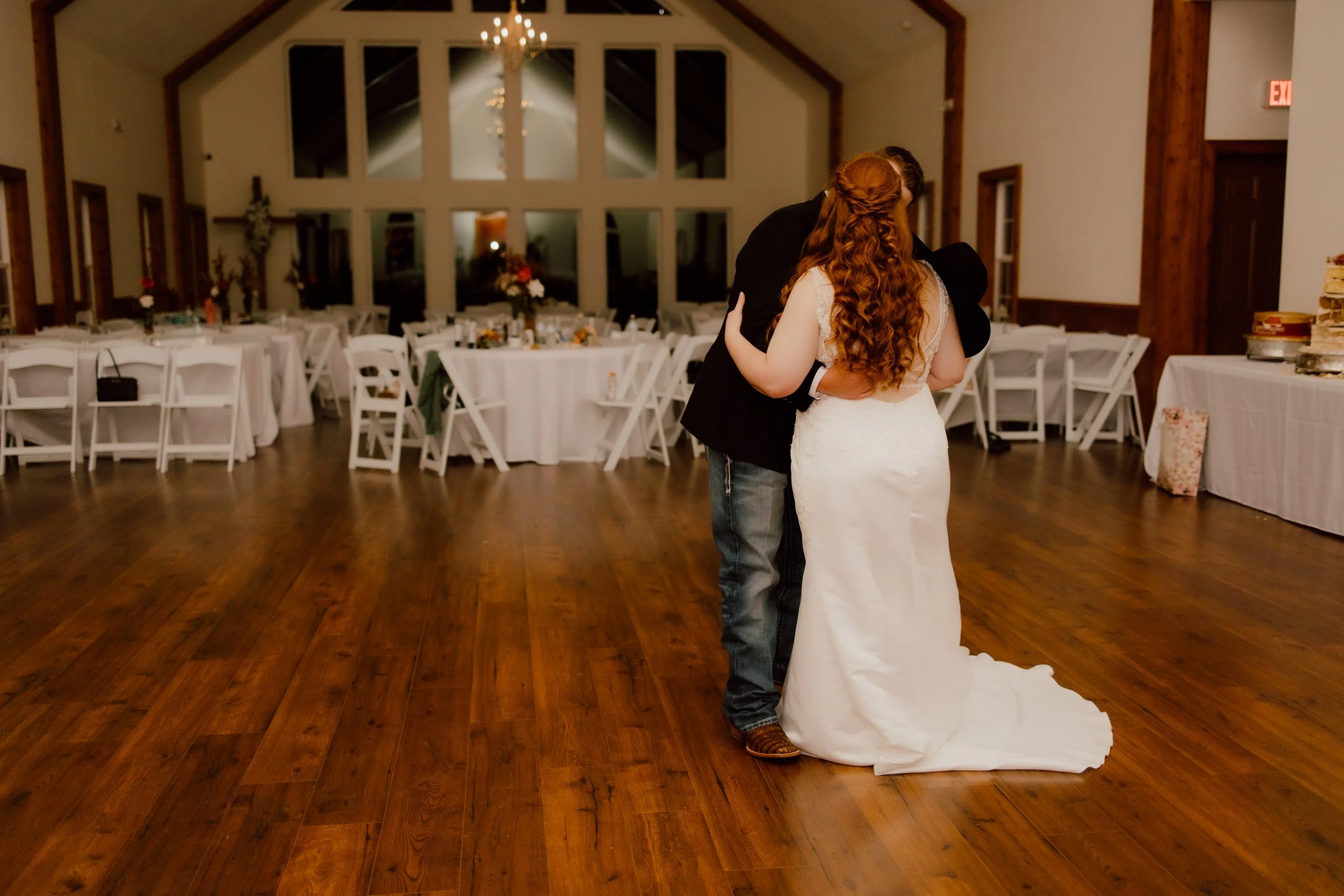 A woman in a white wedding dress and a man in a black suit share a dance in a reception hall decorated with white tables and chairs and floral centerpieces, while embracing.