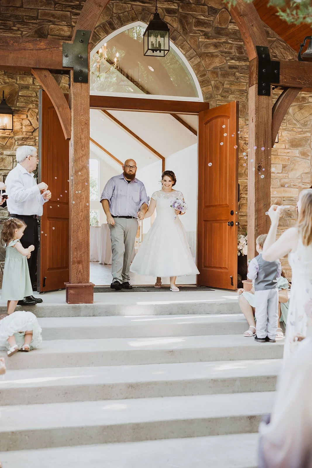 Bride and groom walking out of a rustic stone chapel with open wooden doors, surrounded by friends and family, during a wedding celebration.
