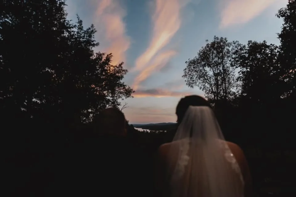 Back view of a bride wearing a veil, standing outdoors at sunset, with trees on either side and a colorful sky above.