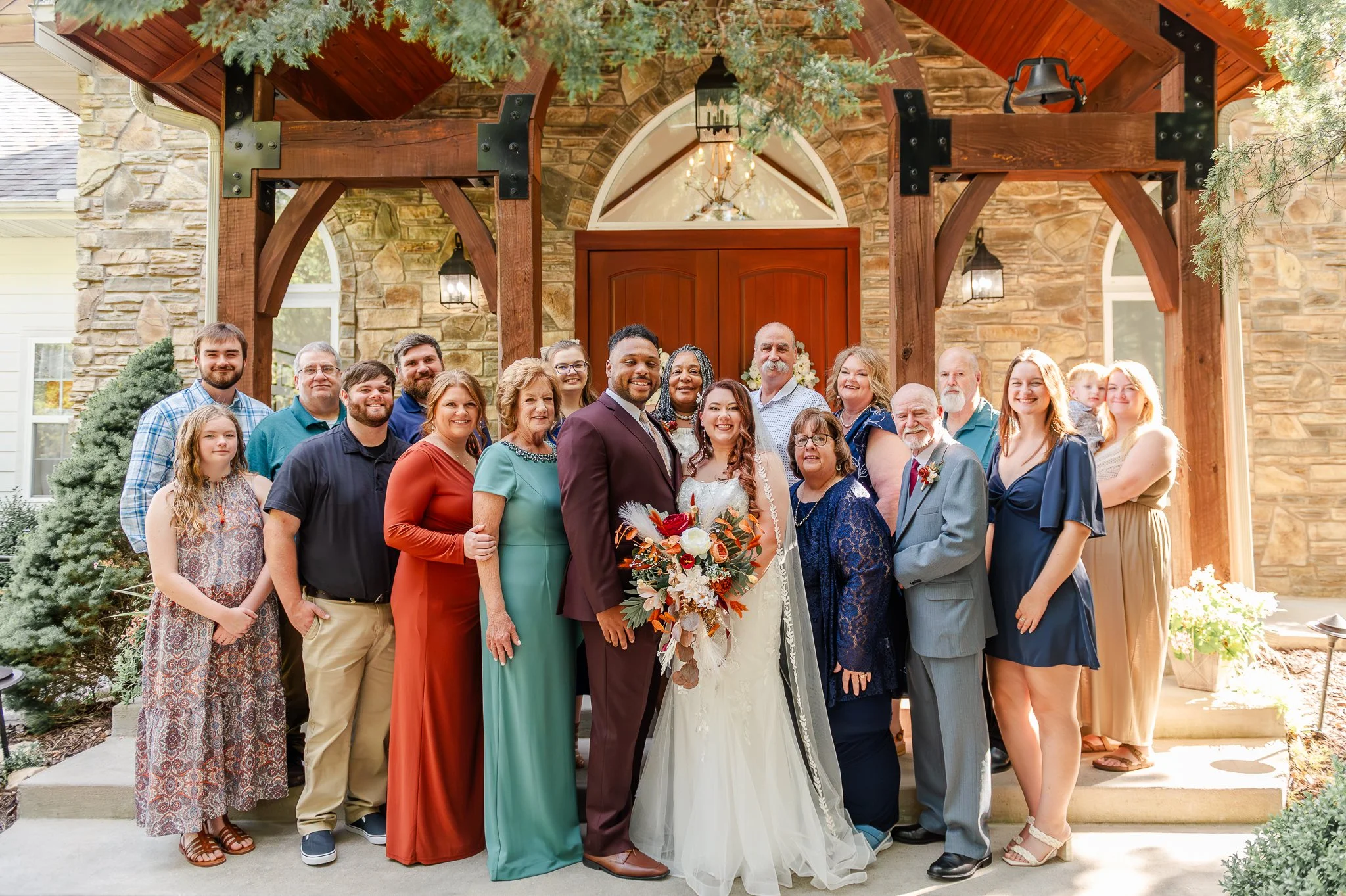 A diverse group of people gathered outside a stone and wood house, celebrating a wedding. The bride and groom are in the center, smiling, with the bride holding a bouquet of flowers. The group includes men, women, and children of various ages, dressed in semi-formal attire. The house has large wooden beams, a wooden door, and hanging lanterns. Pine trees frame the scene.