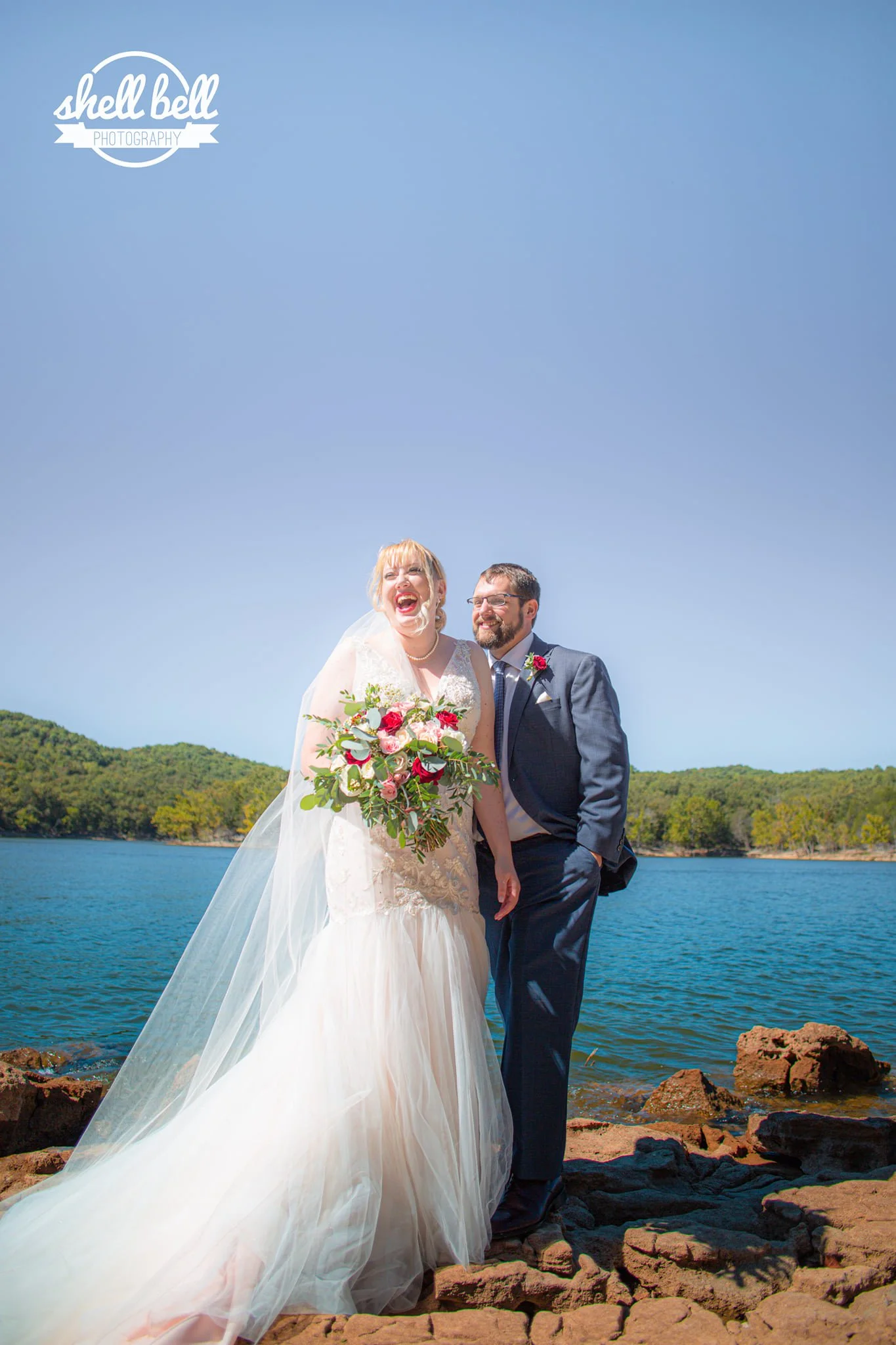 A bride and groom standing on rocks by a lake with trees and hills in the background, smiling and celebrating their wedding on a sunny day.