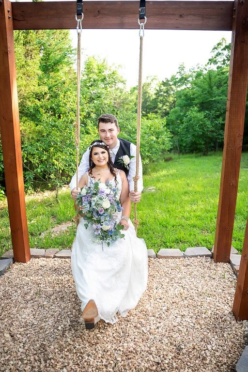 A newlywed couple sitting on a swing in a garden. The bride holds a bouquet of flowers and wears a white wedding dress, while the groom sits behind her, smiling, and is dressed in a black vest and white shirt.