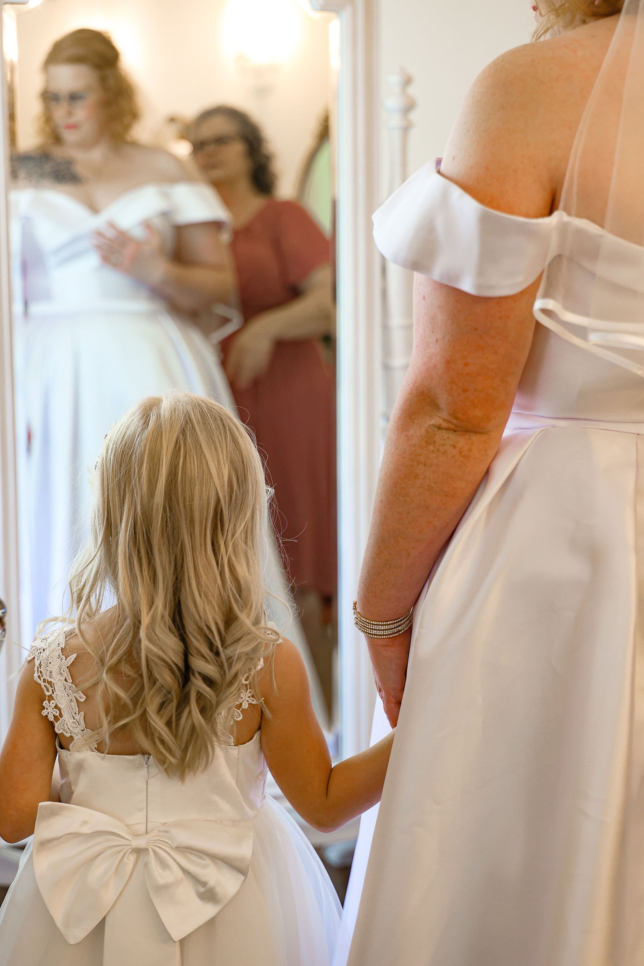 A young girl with long blonde hair and a white dress with lace details on the sleeves, looking at a woman in a white dress, likely a bride, with her back to the camera. The bride's dress has off-the-shoulder sleeves and a large bow at the waist. Two women are reflected in a mirror, one wearing glasses and a reddish dress, and the other with curly hair and glasses. The scene appears to be in a dressing room or bridal suite.