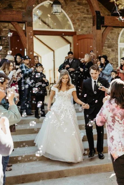 Bride and groom walking down the stairs holding hands at a wedding reception, surrounded by guests, with bubbles in the air.