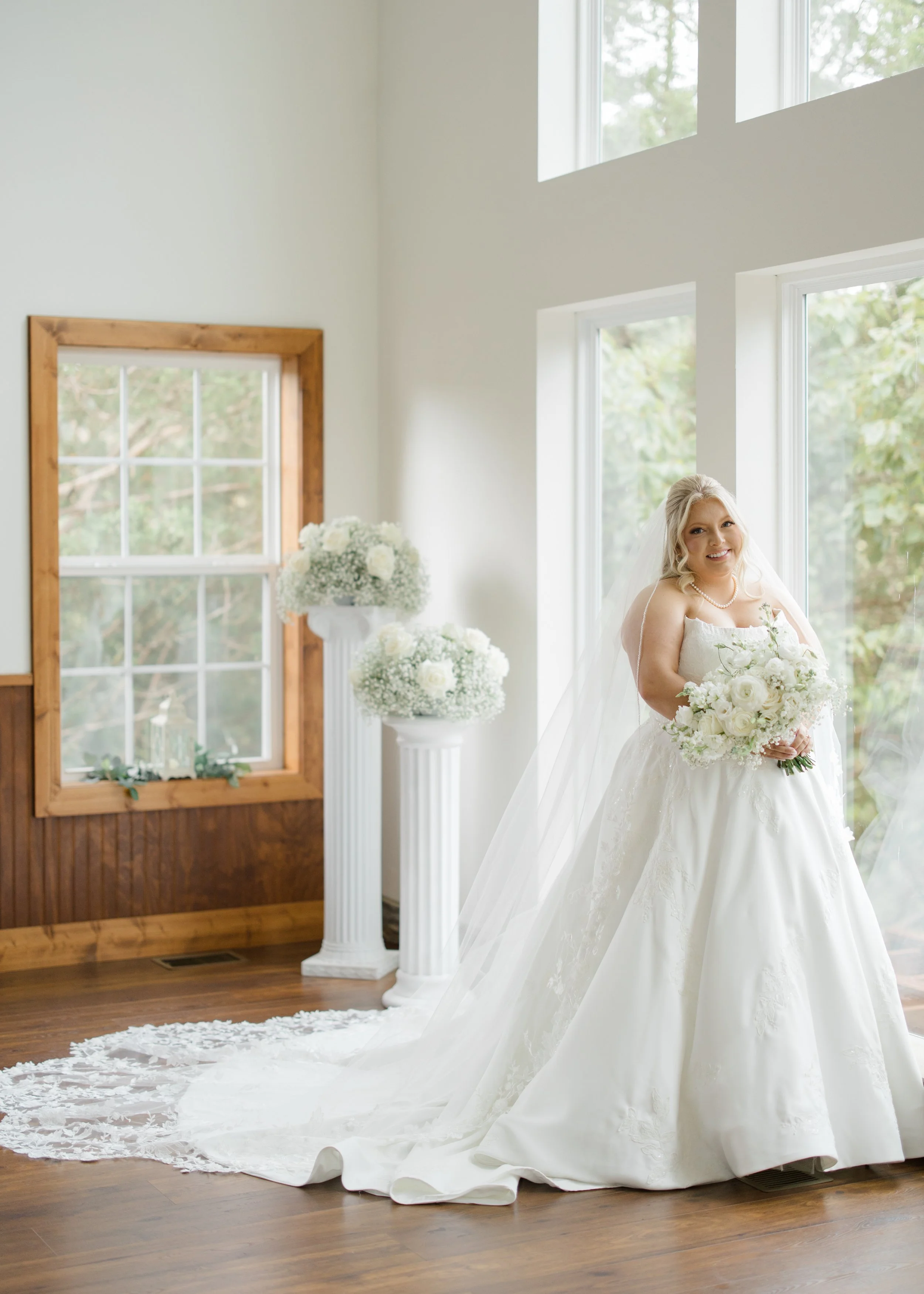 A bride in a white wedding gown holding a bouquet of white flowers, standing indoors near large windows with a wooded view outside, in a room decorated with white flowers and wooden accents.