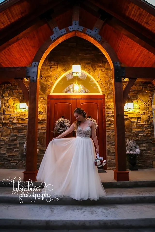 Bride in a wedding dress standing on steps outside a stone and wood building, holding a bouquet, under a lit archway.