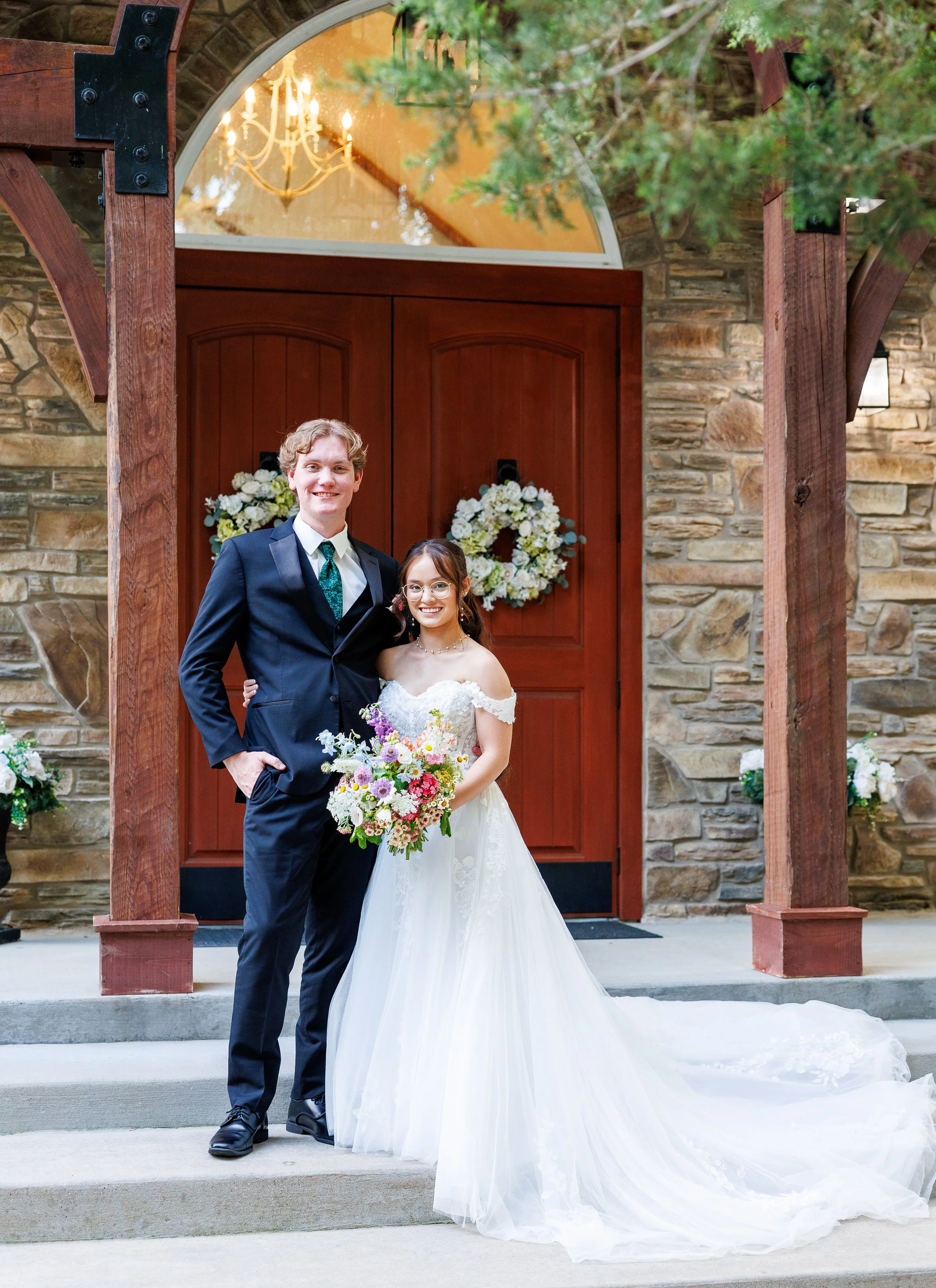 A newly married couple standing together on the steps of a building with a stone wall and wooden door, decorated with white floral wreaths, with the bride holding a colorful bouquet and wearing a white wedding dress, and the groom in a dark suit with