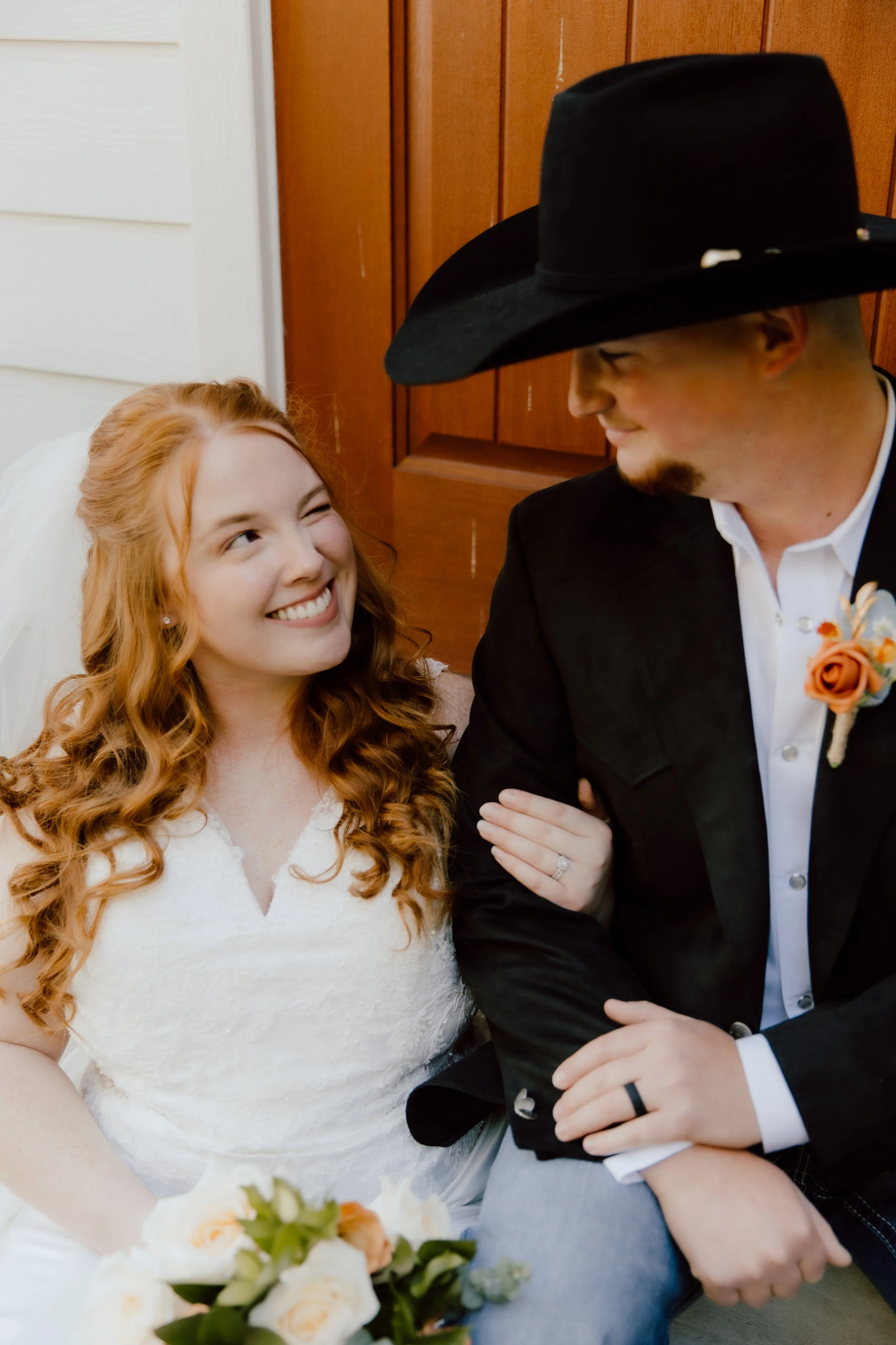 A bride and groom are smiling and looking at each other during their wedding. The bride has long curly auburn hair and is wearing a white lace dress, holding a bouquet of white and peach flowers. The groom is wearing a black suit with a white shirt, 